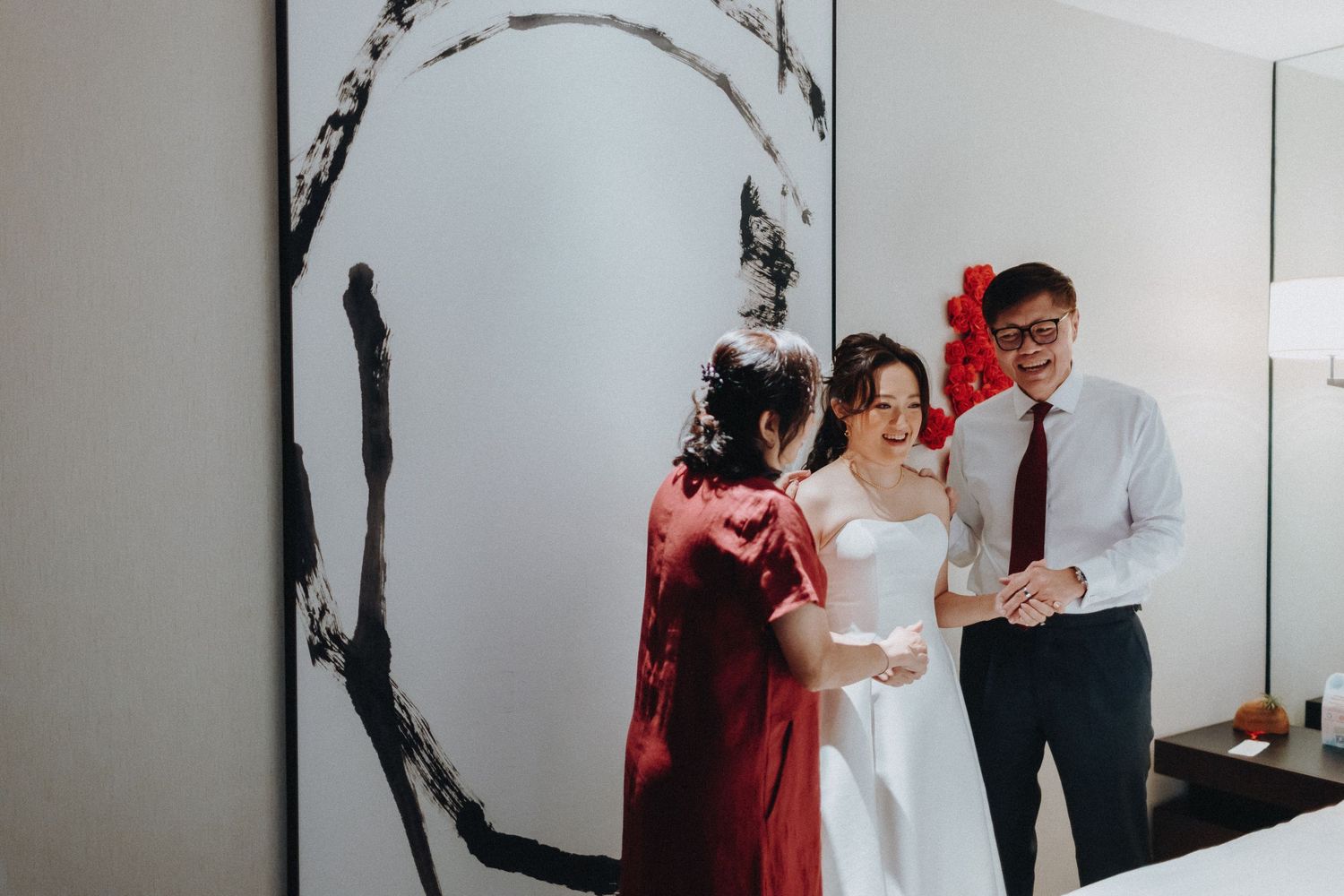 Wedding party sharing joyful candid moments together against a minimalist white wall with red floral decor.