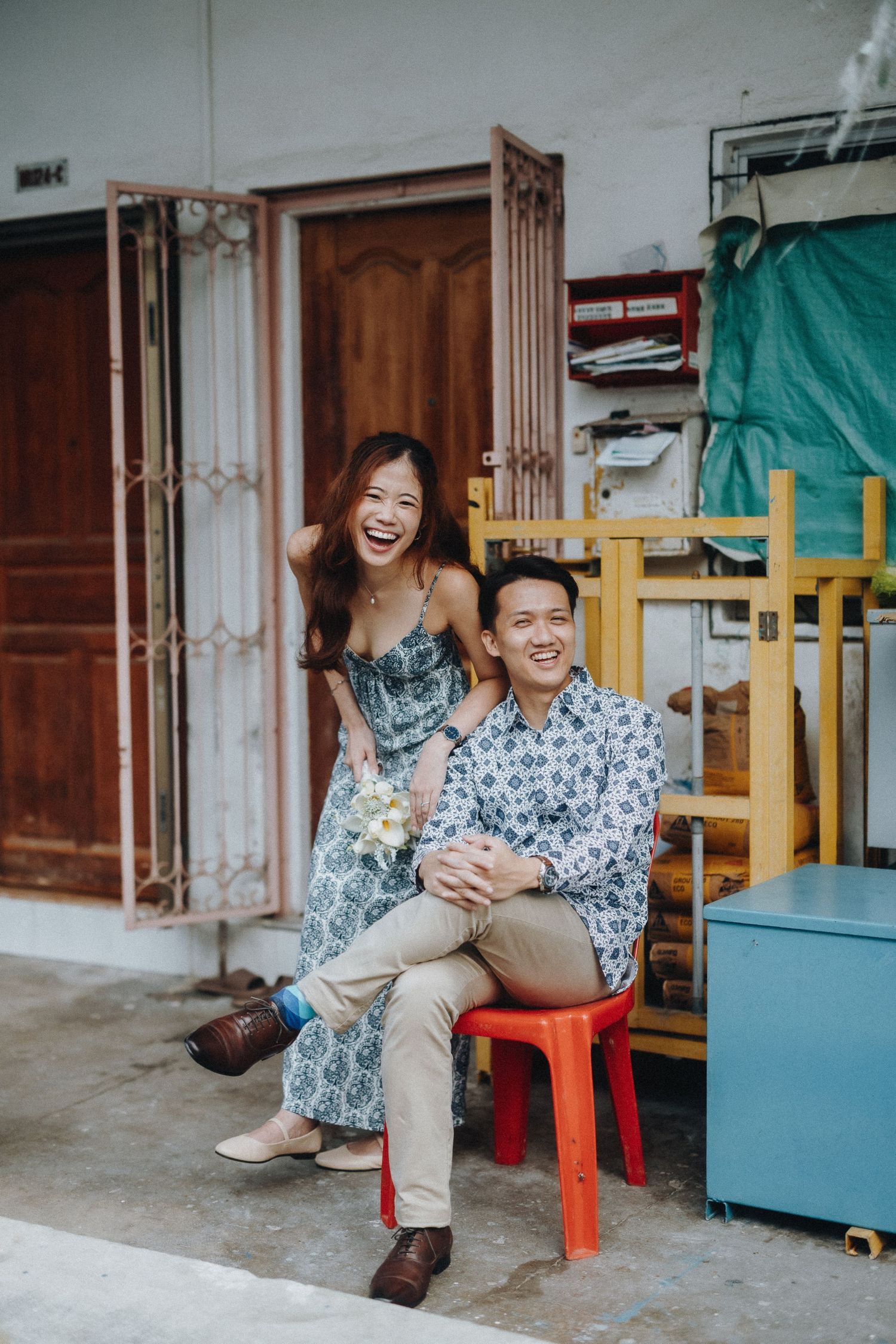 Two people share genuine laughter while sitting on a bright red stool outside a building entrance.