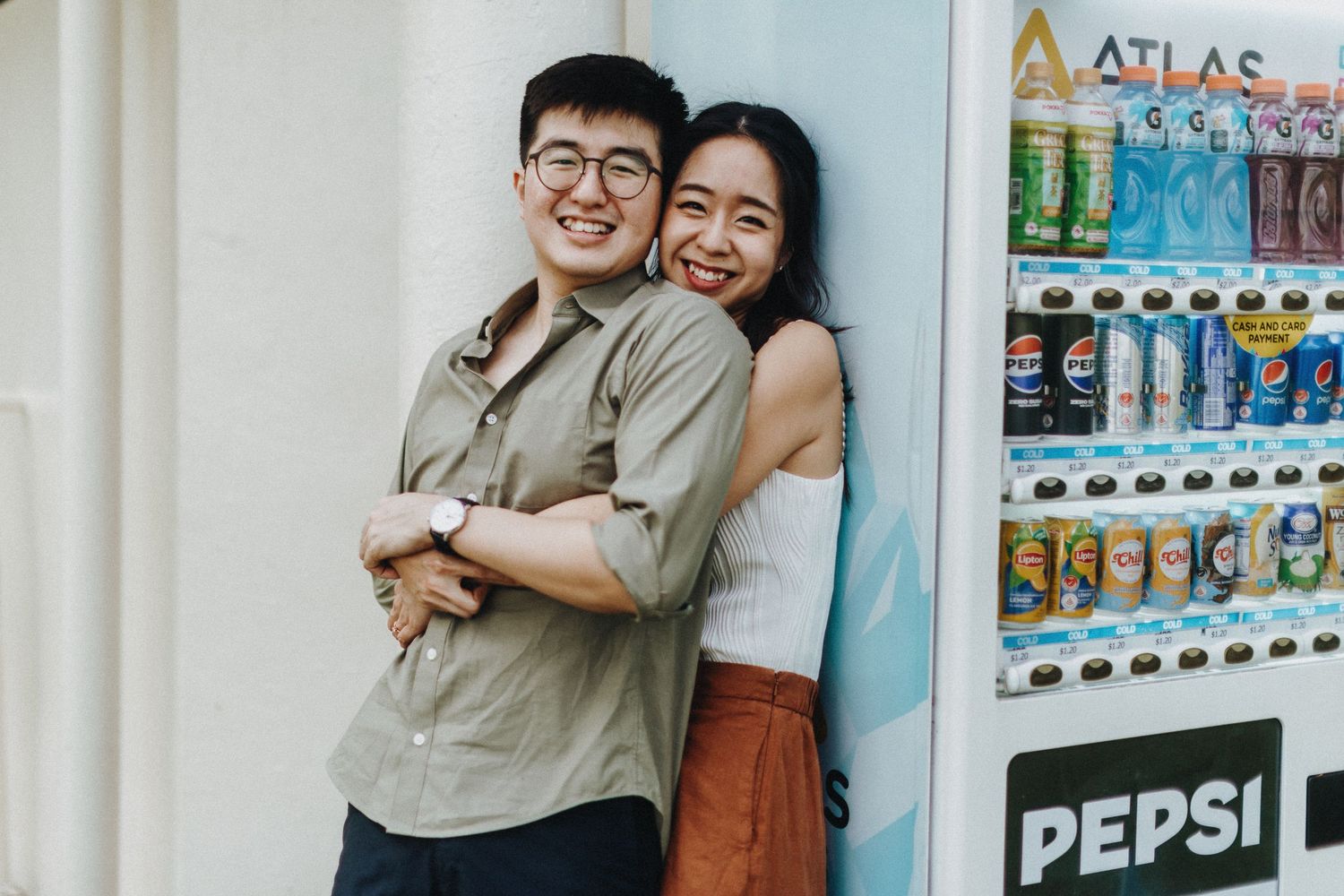 A couple embraces and smiles in front of a vending machine filled with colorful beverages and snacks.