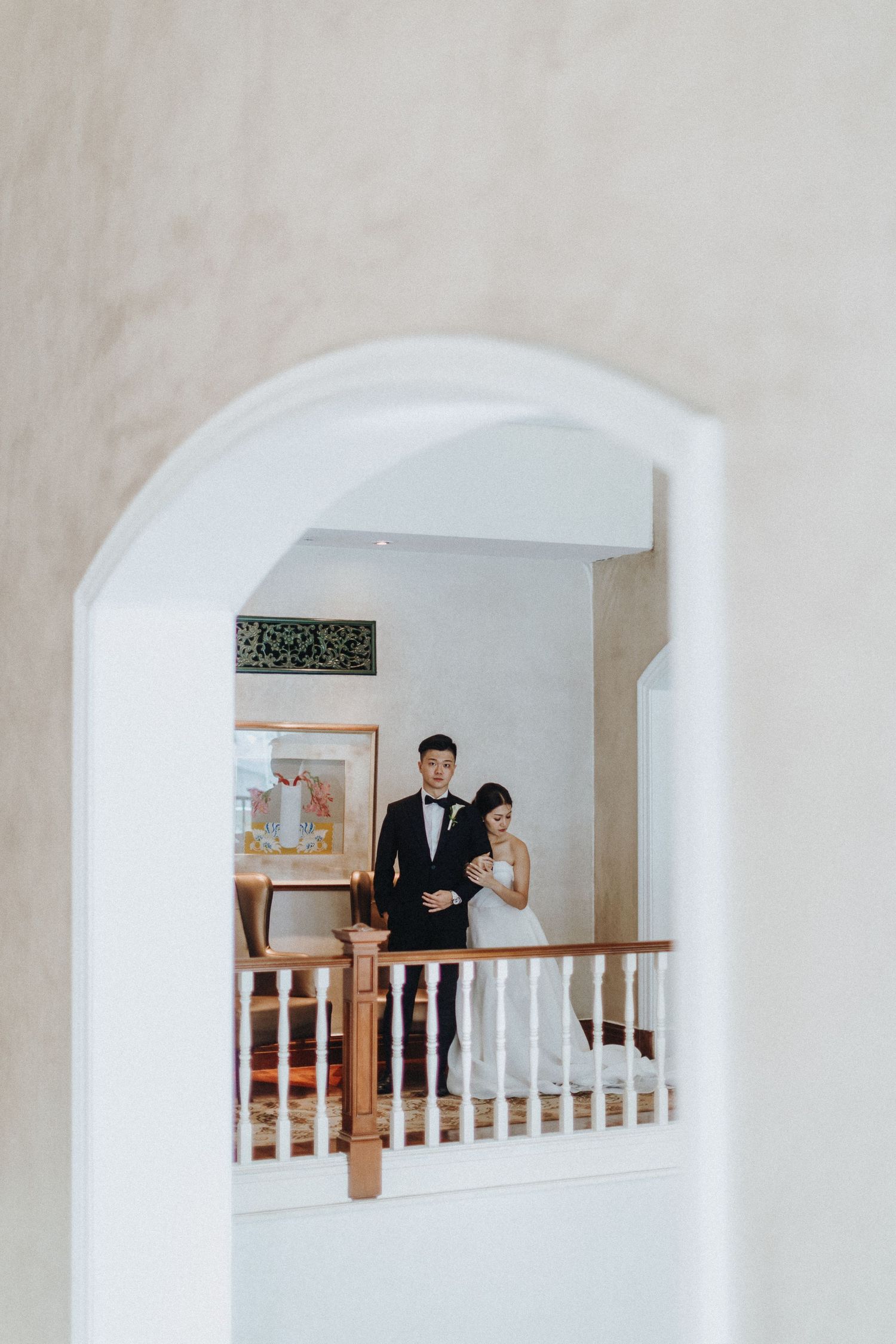 Couple in formal wedding attire standing behind white archway and banister in an elegant indoor setting.