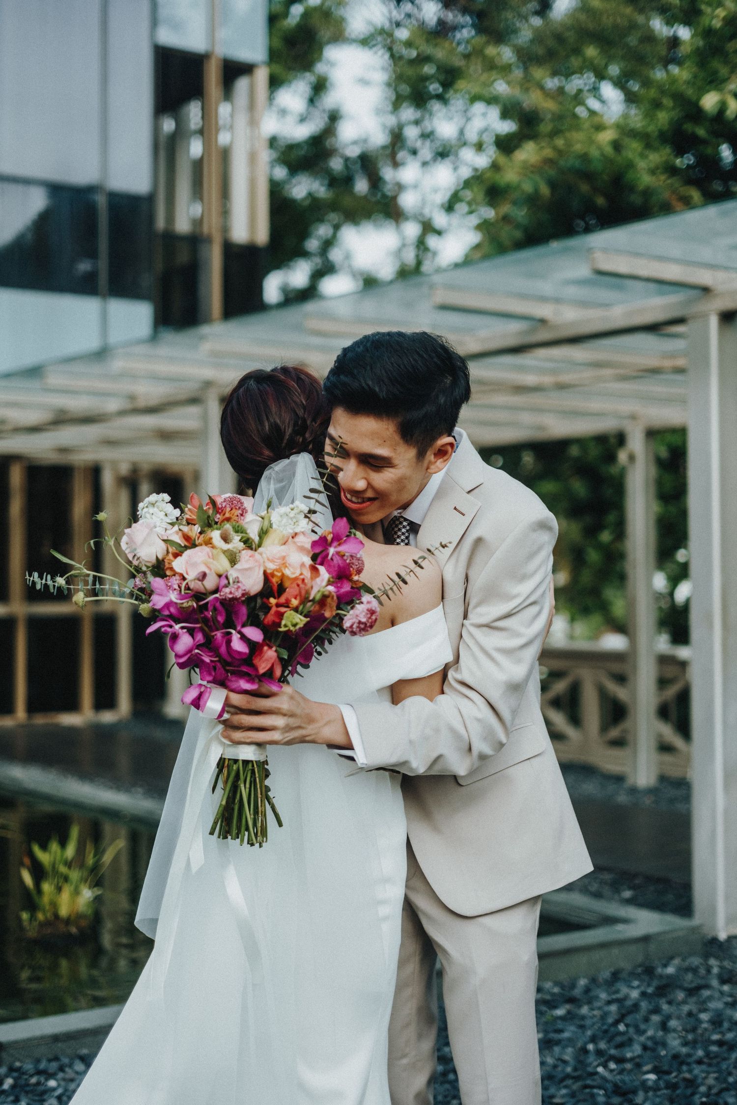 A couple in wedding attire share a moment with vibrant pink and purple floral bouquets outdoors.