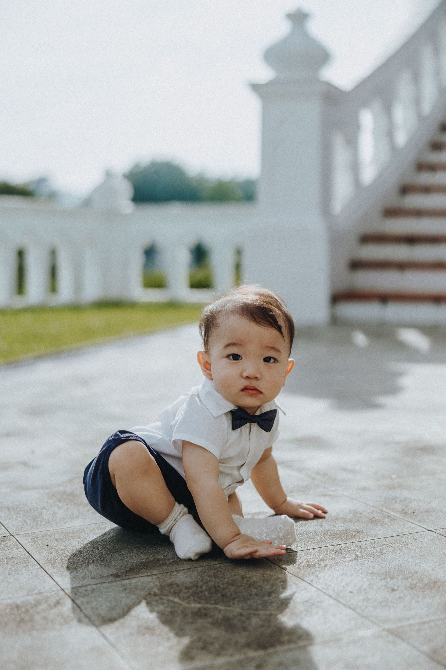 A toddler in formal wear with bow tie crawls on a marble floor near white pillars and steps.