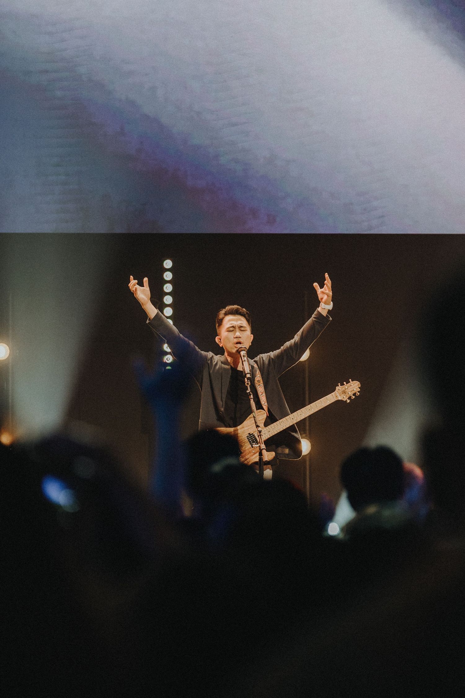 A musician performs energetically on stage with raised arms while holding a guitar in front of a dark crowd.