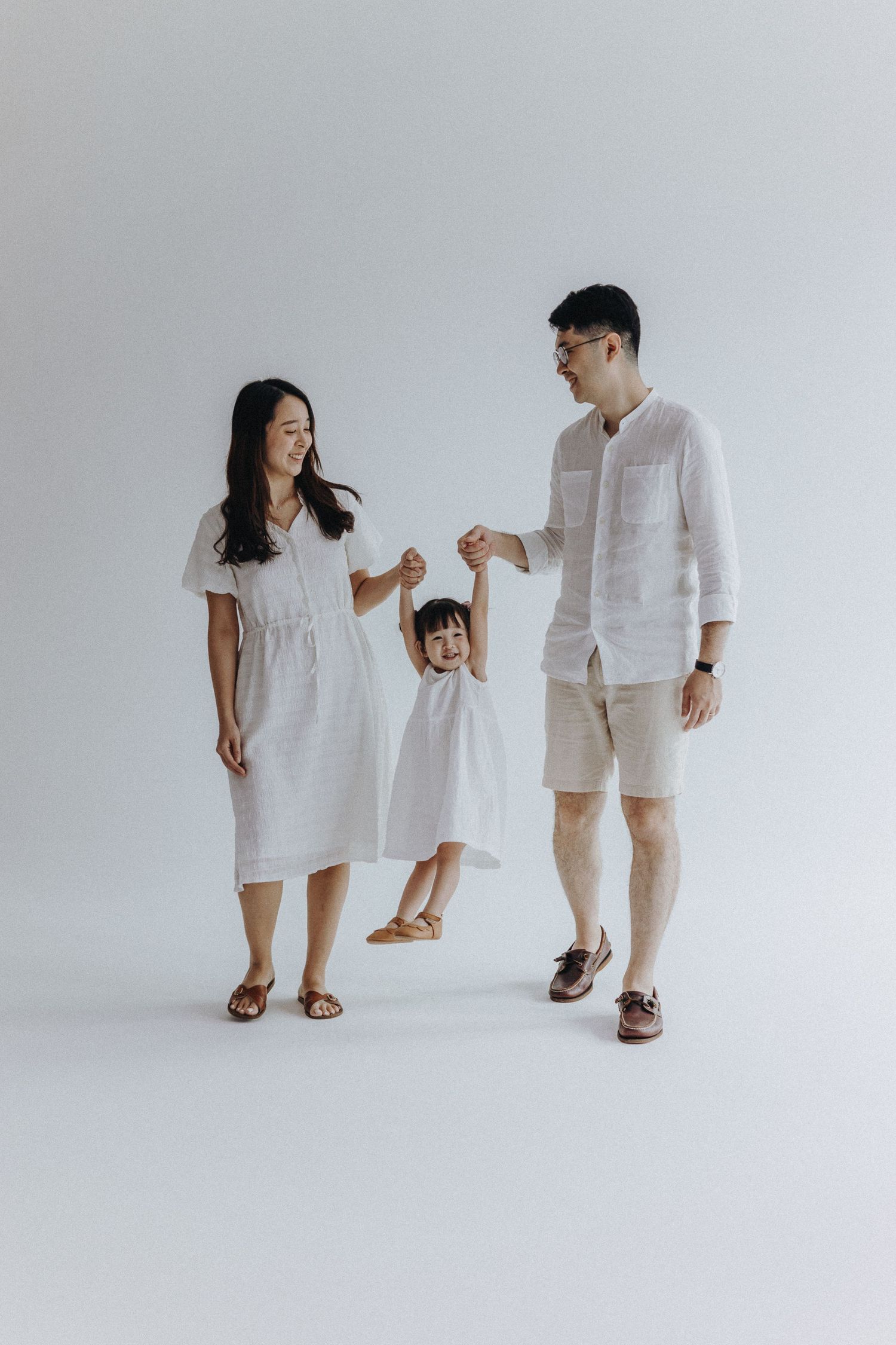 Family wearing white outfits poses playfully in minimalist studio setting on light background.