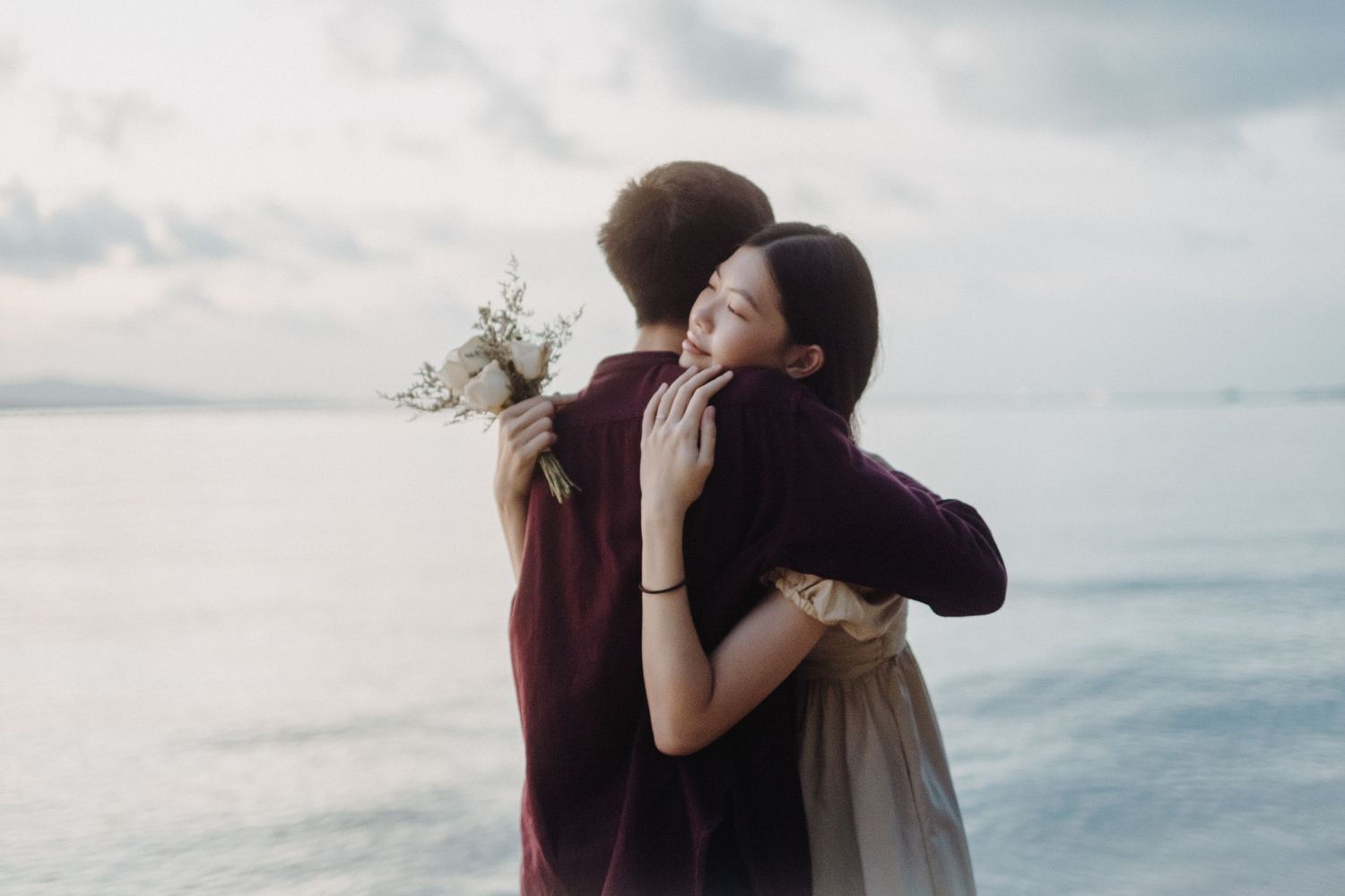 Two people embrace in a tender moment at sunset near the ocean, their silhouettes casting long shadows on the beach.
