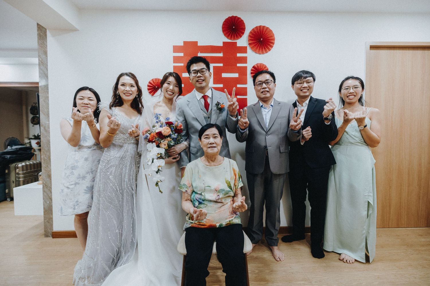 A traditional Chinese wedding celebration indoors with attendees in formal attire standing together against a wall with red decor.