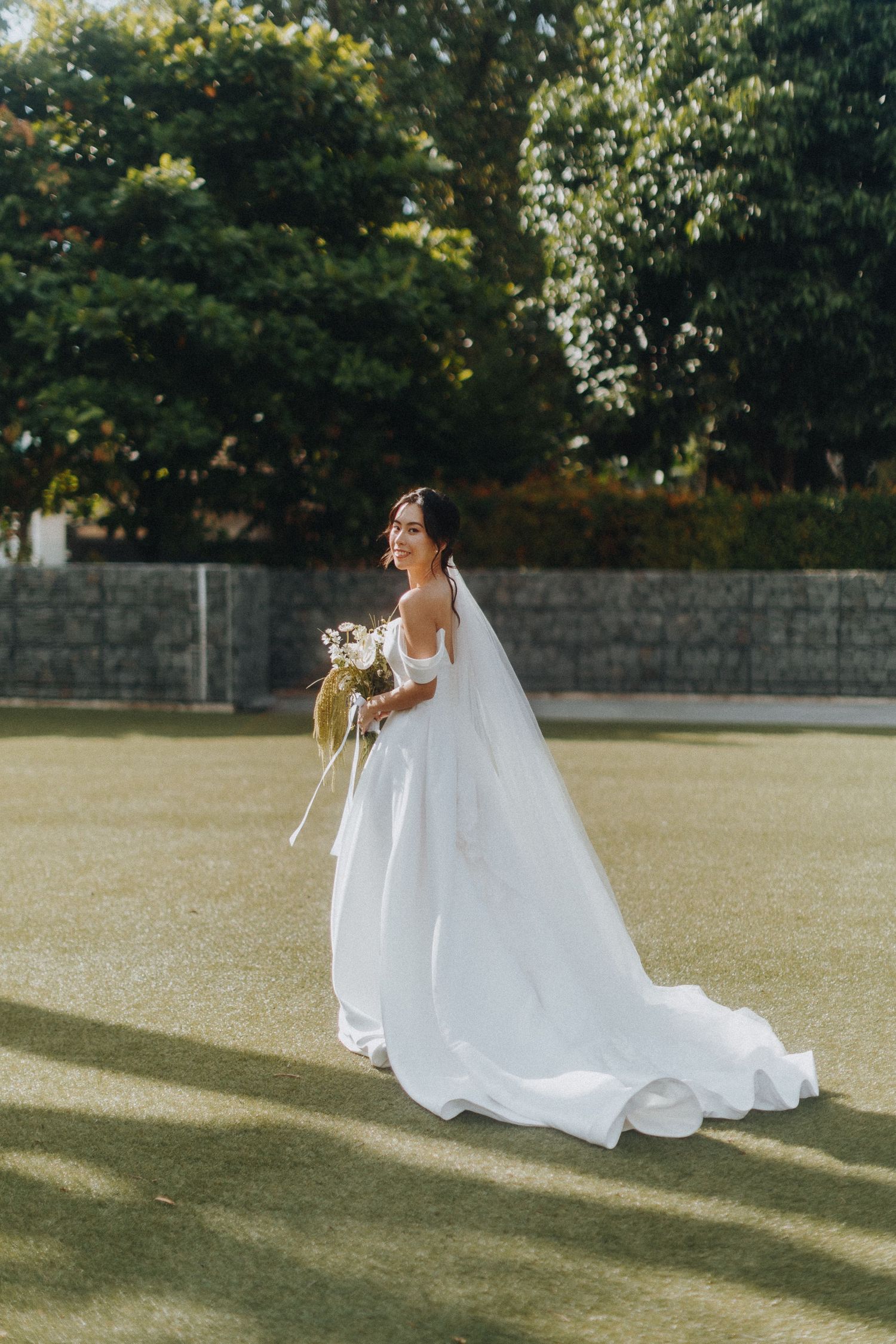 A wedding bride in a flowing white gown with long train poses in a sunlit garden setting holding a bouquet.