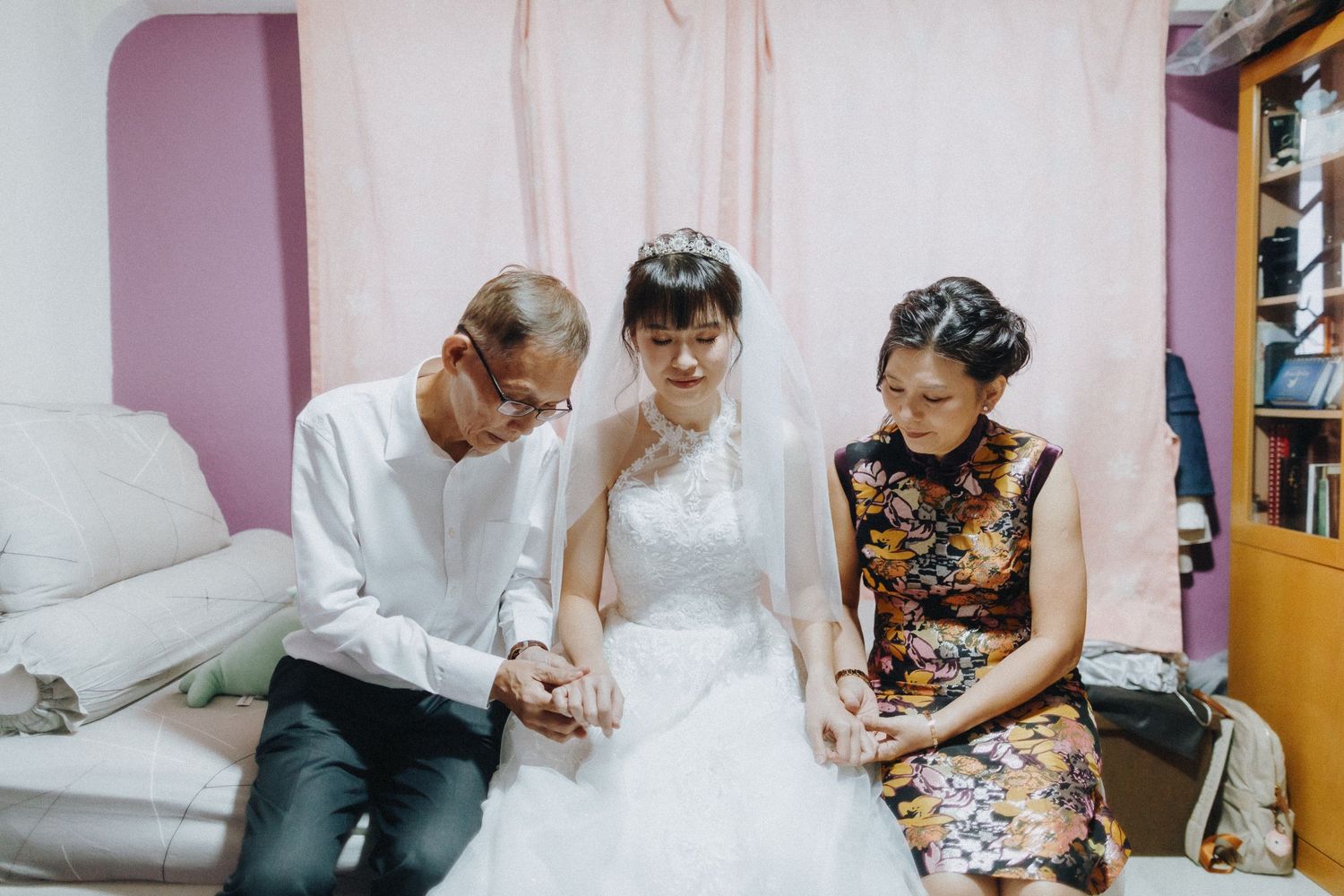 A quiet moment shared during wedding preparations in a pink-walled room with a bridal gown and floral dress.