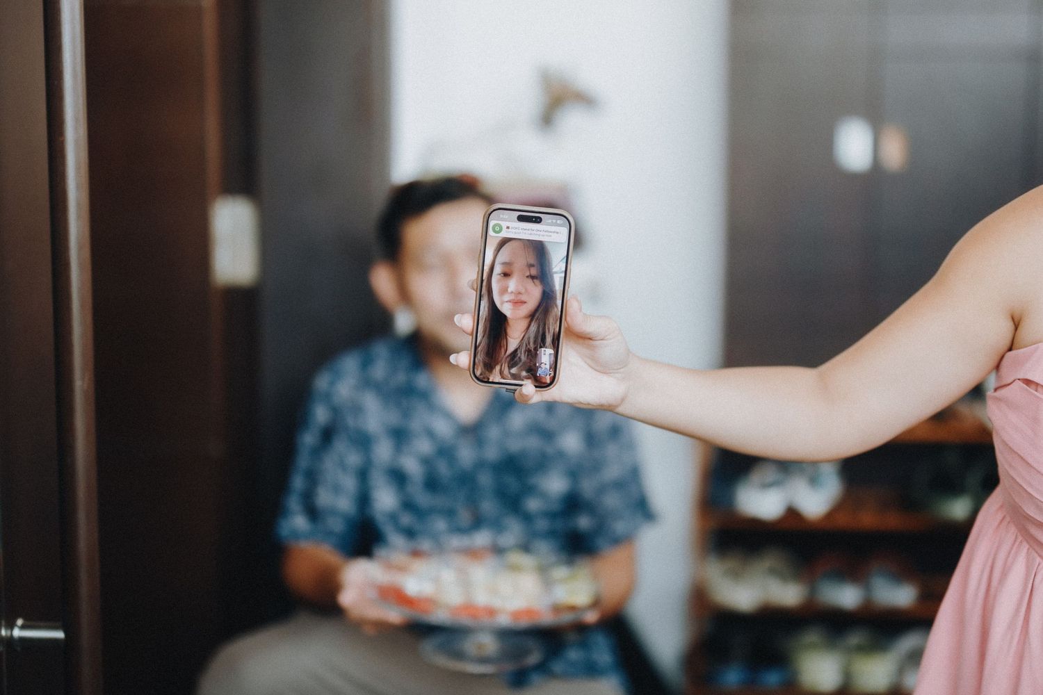 A hand holds up a smartphone taking a selfie photo while someone sits in the background with food.