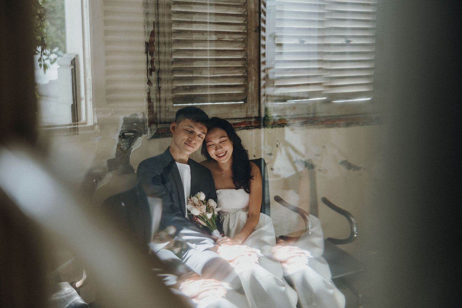 A couple in wedding attire rests against a wall with window blinds in a soft, intimate moment.