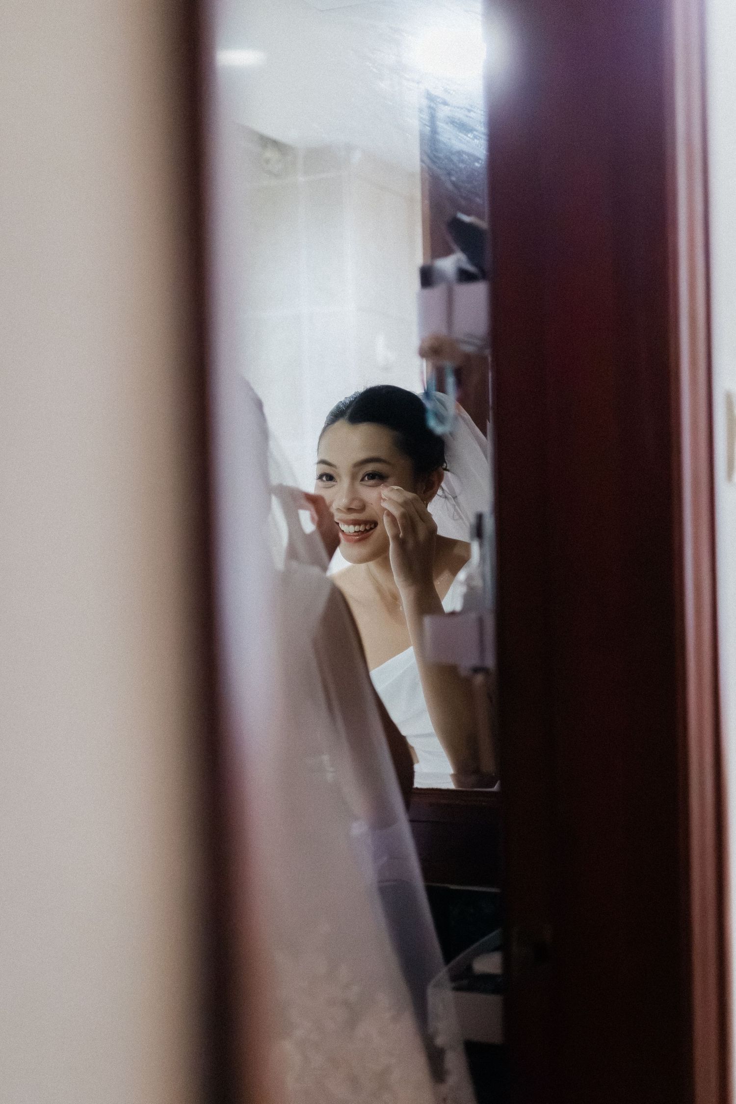 Bride applying makeup while smiling at her reflection in a mirror on her wedding day.