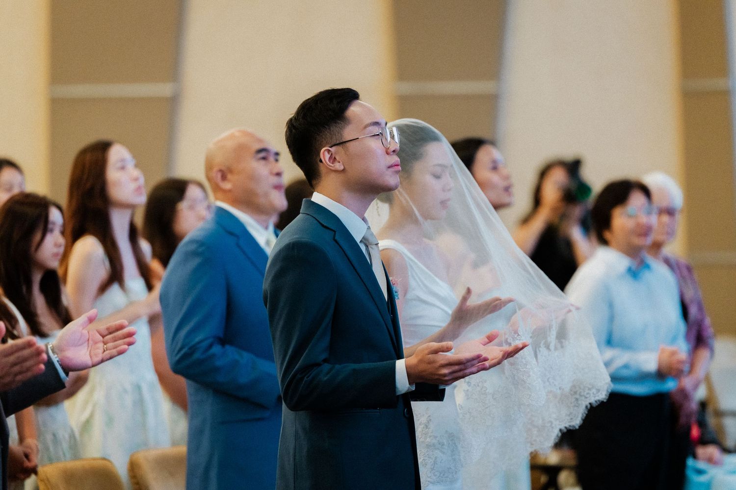 Asian couple in wedding attire clapping during church ceremony surrounded by guests in formal wear.
