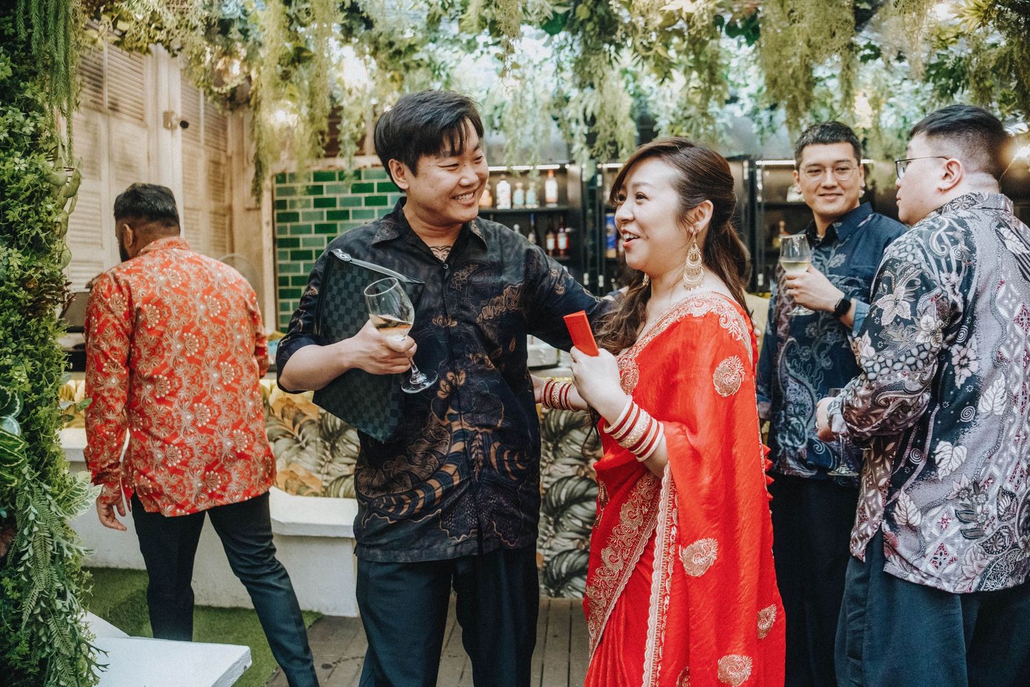 Guests in traditional attire toast with wine glasses at an elegant indoor garden wedding reception.
