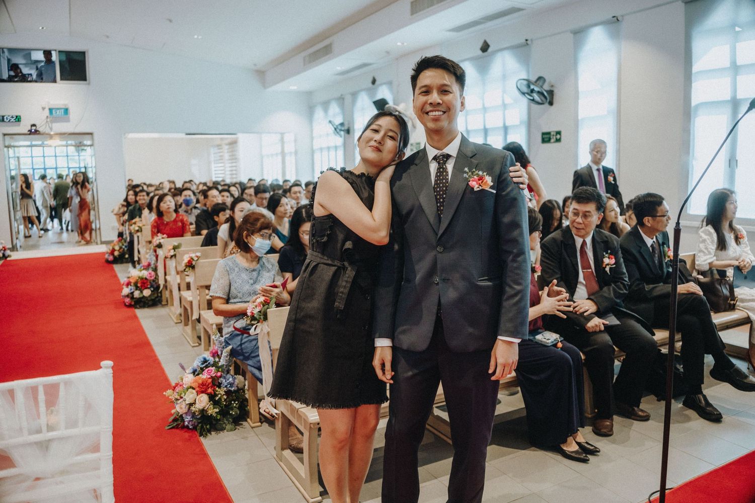 Asian couple posing together in a bright church wedding venue with guests seated along a red carpet aisle.