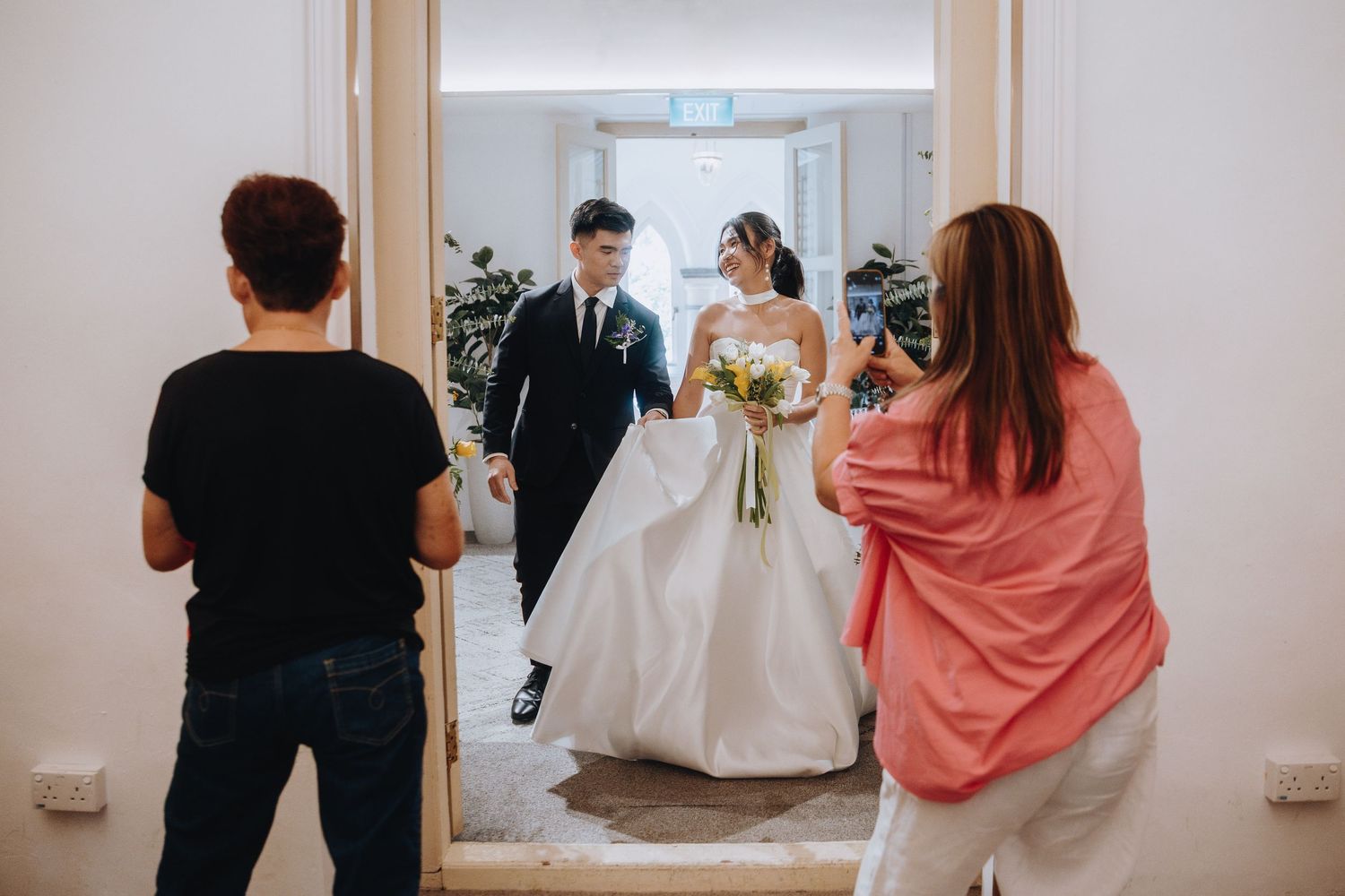 Wedding photographers capture bride and groom entering through a doorway in a bright elegant venue.