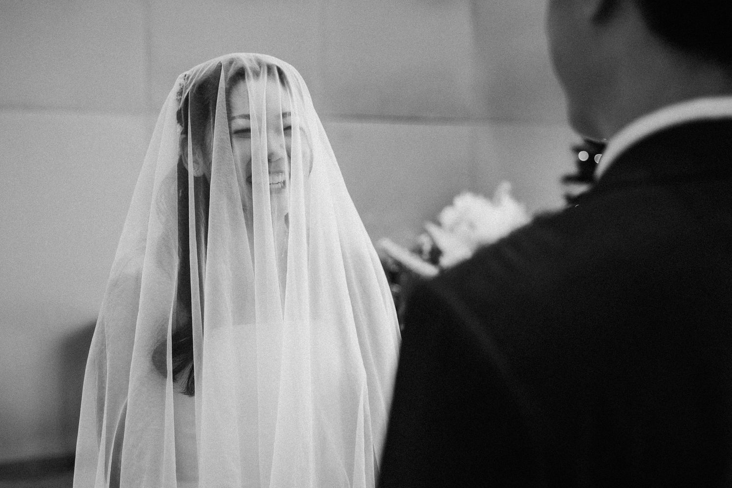 Beautiful veiled bride smiles softly at groom during emotional black and white wedding ceremony.