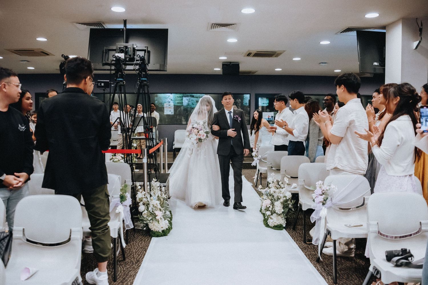 Wedding ceremony in progress with white aisle runner and floral decorations in an indoor venue.