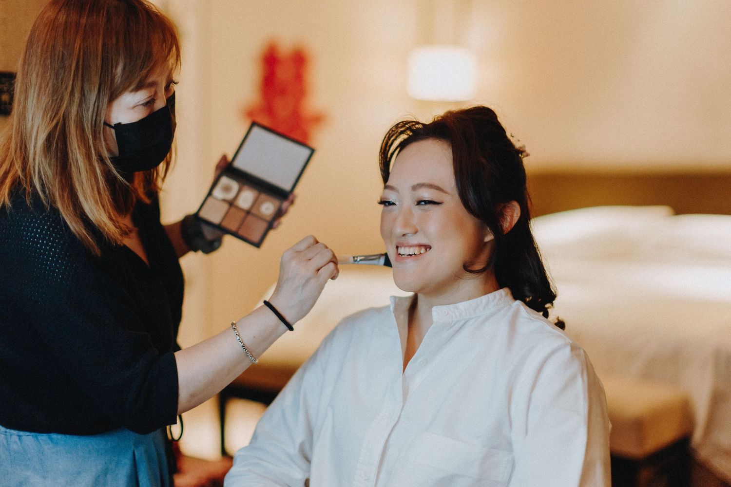 Bride getting makeup applied while sitting in a white robe during wedding preparations.