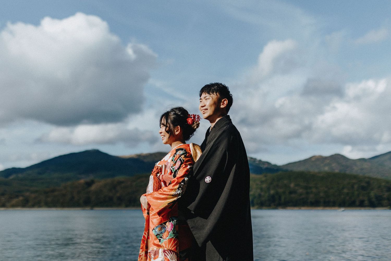 A couple in formal attire stands at the edge of a lake with mountains and dramatic clouds in the background.