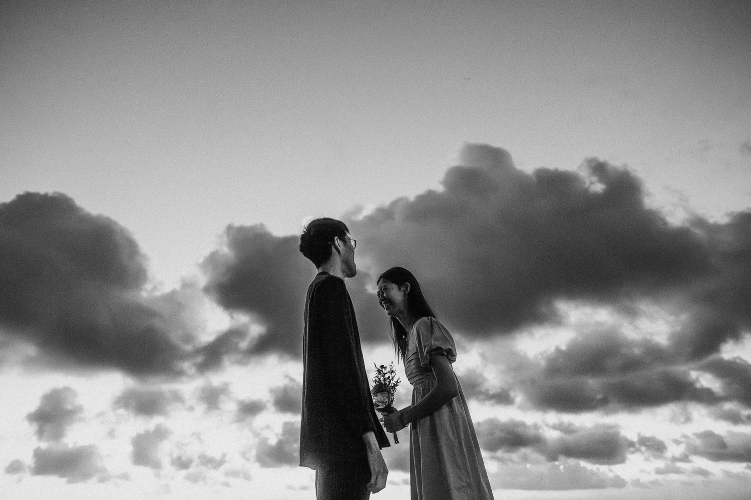 Silhouette of a tree and couple against dramatic cloudy sunset sky creating romantic black and white composition.
