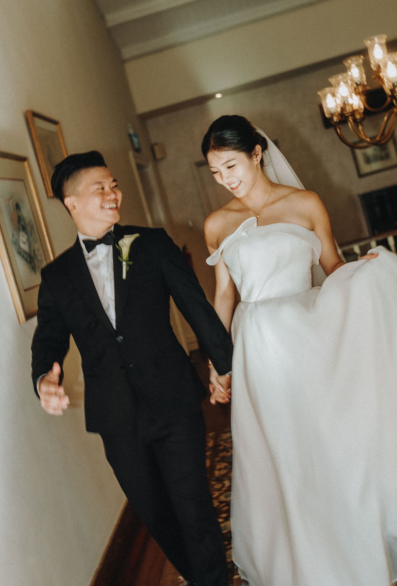 A couple in formal wedding attire walking together and holding hands in an elegant indoor setting.