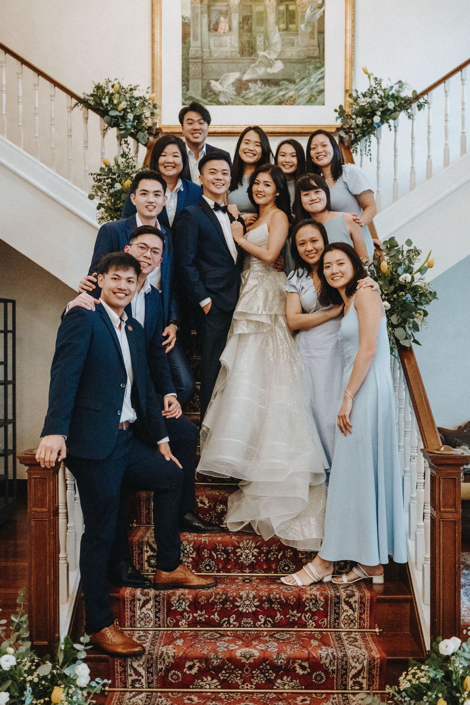 Wedding party poses together on stairs with floral decorations and an ornate carpet runner.