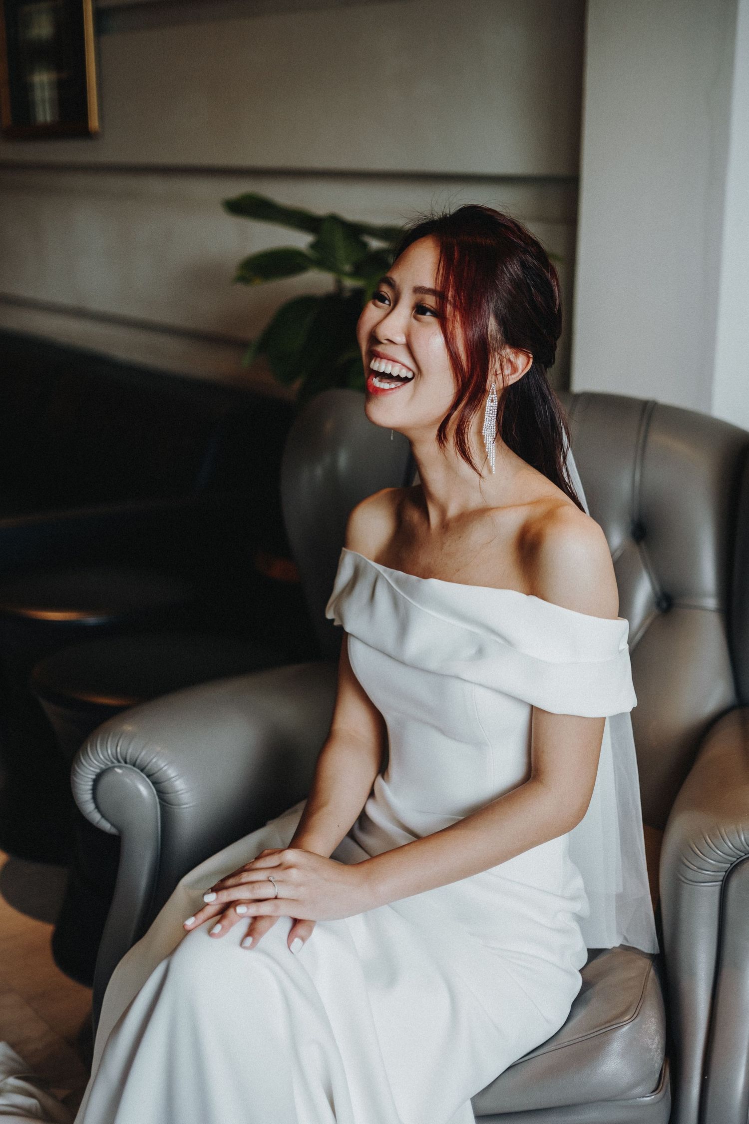 A person in a white off-shoulder dress sits on a grey couch laughing joyfully during a photoshoot.