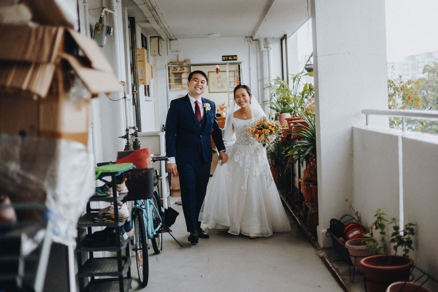 A wedding couple walking through an apartment corridor filled with plants and everyday items.