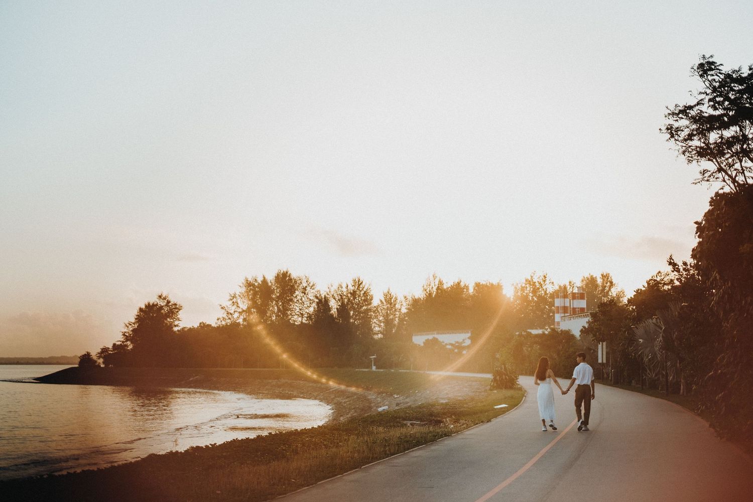 A couple walking along a coastal path during golden hour with sunbeams streaming through trees.