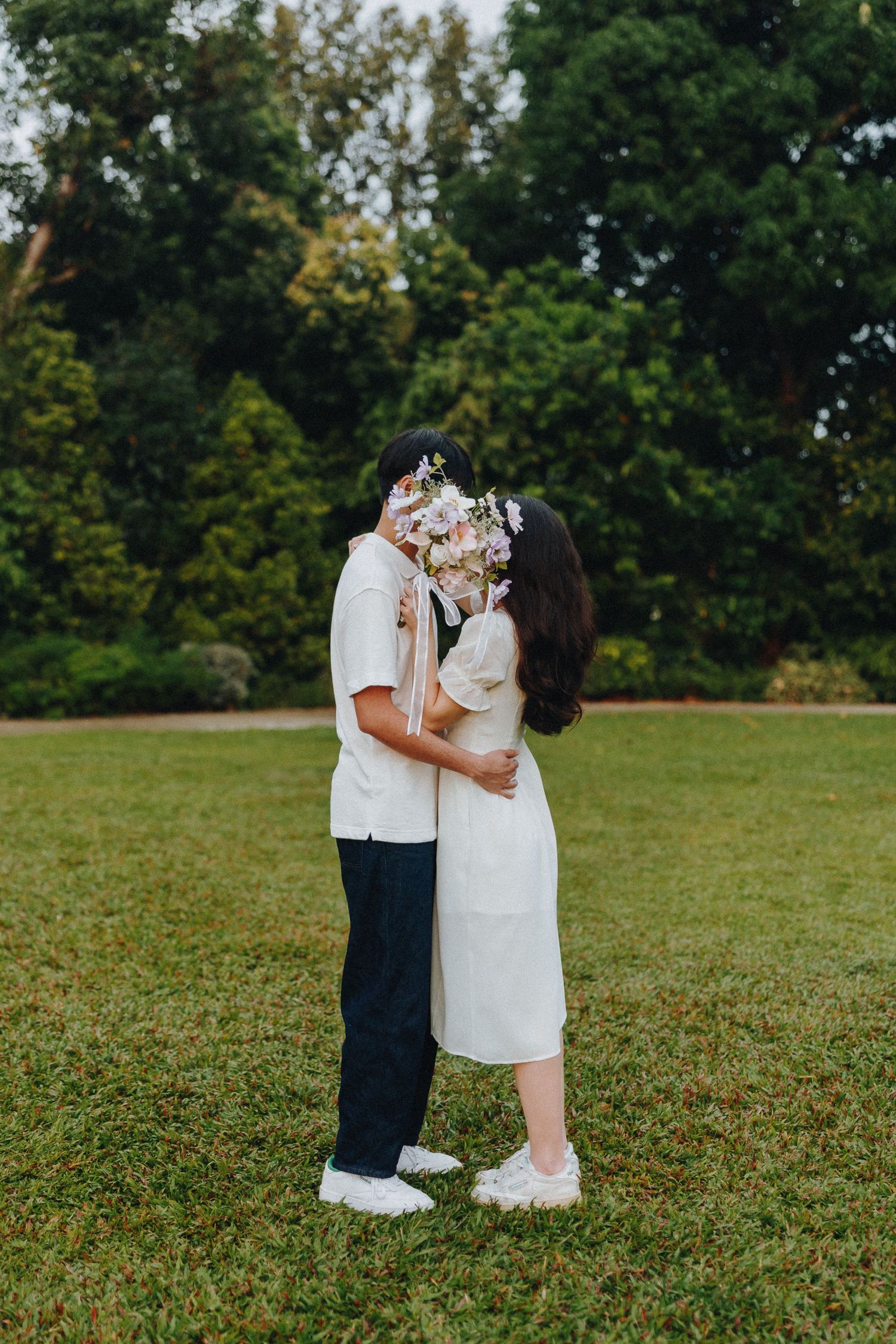 A romantic couple embraces on a grassy lawn while holding a bouquet of flowers.