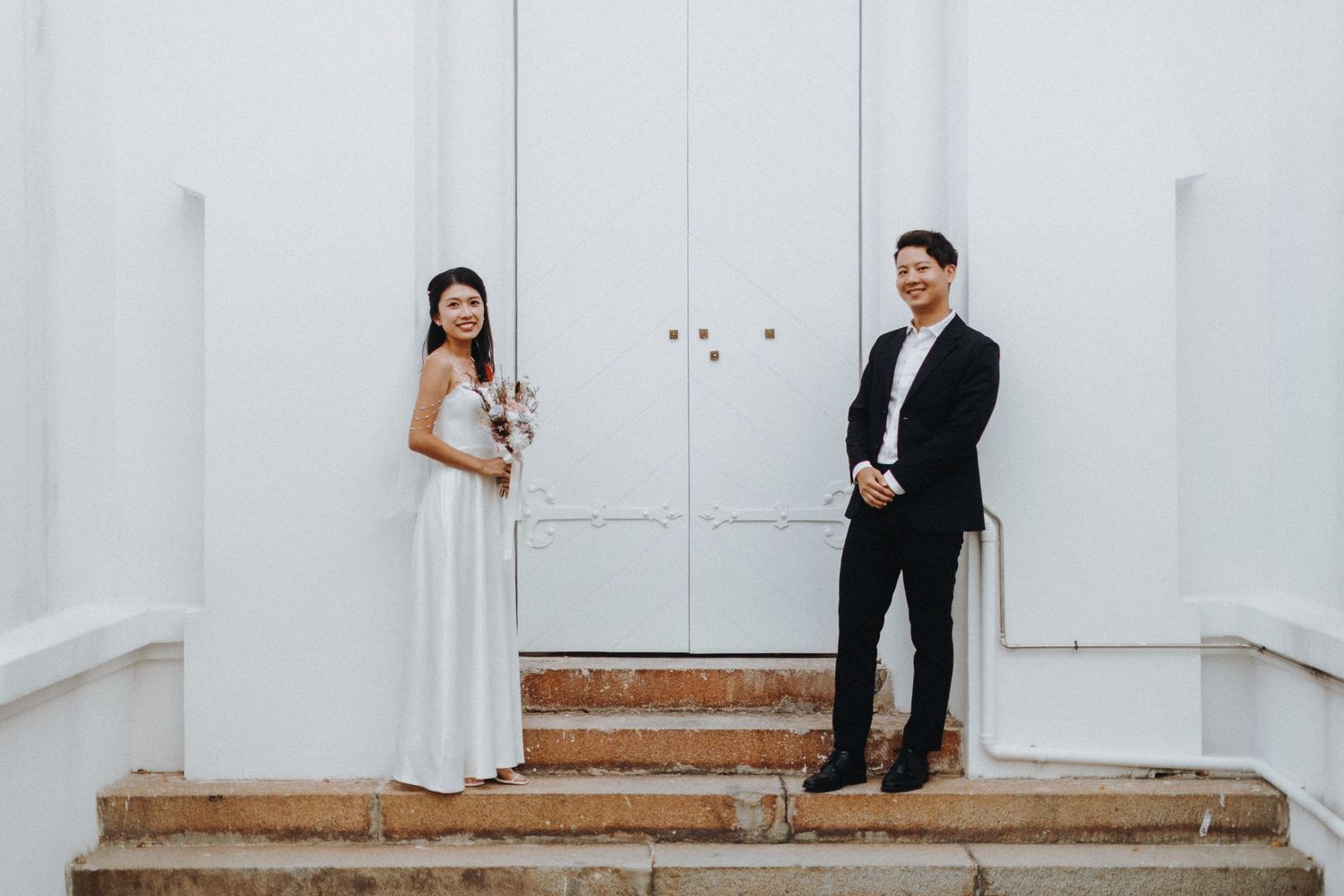 A formal couple poses on white steps against white doors in an elegant setting.