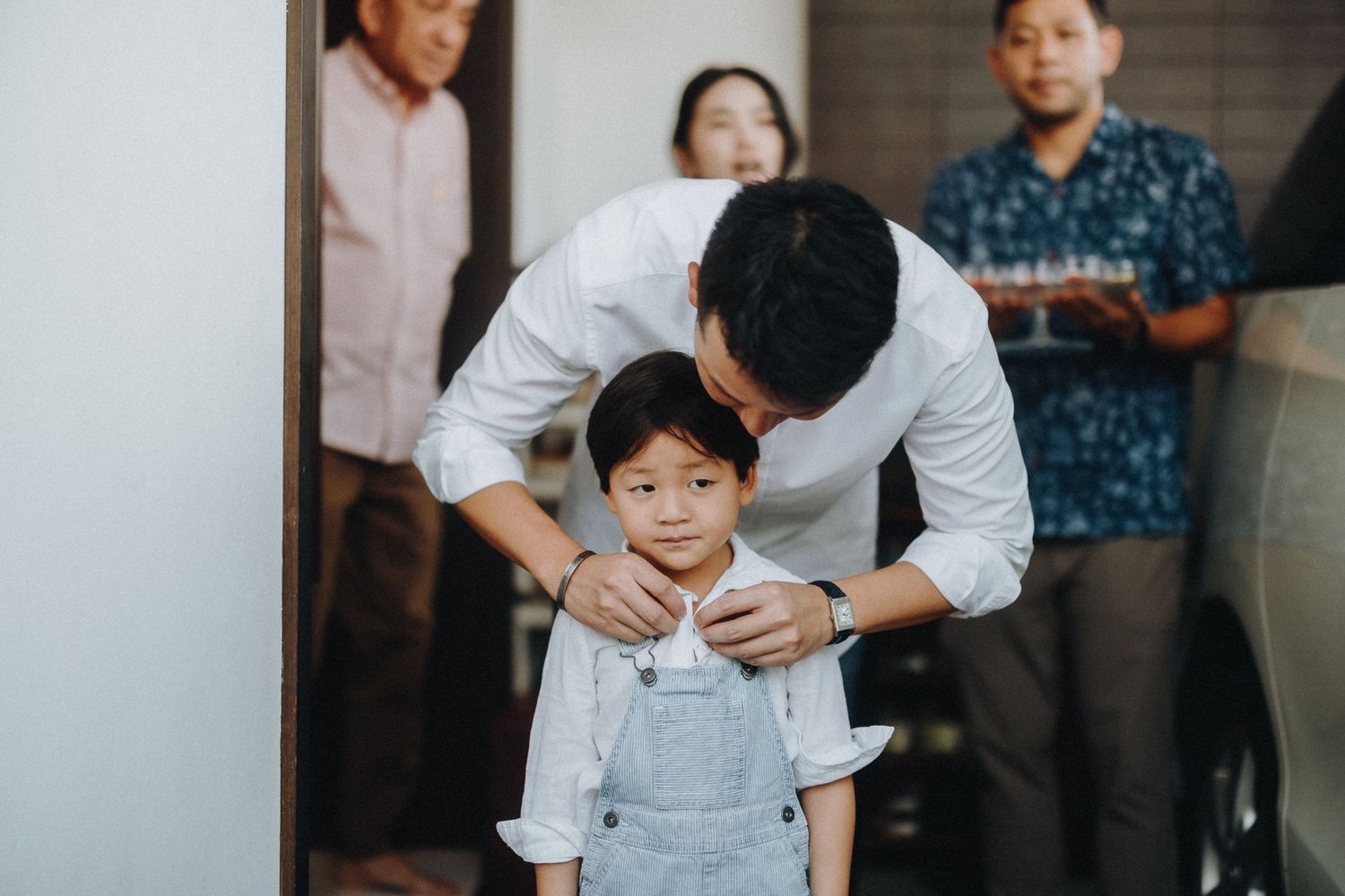 A parent adjusts their young child's clothing while standing in a room with others watching in the background.