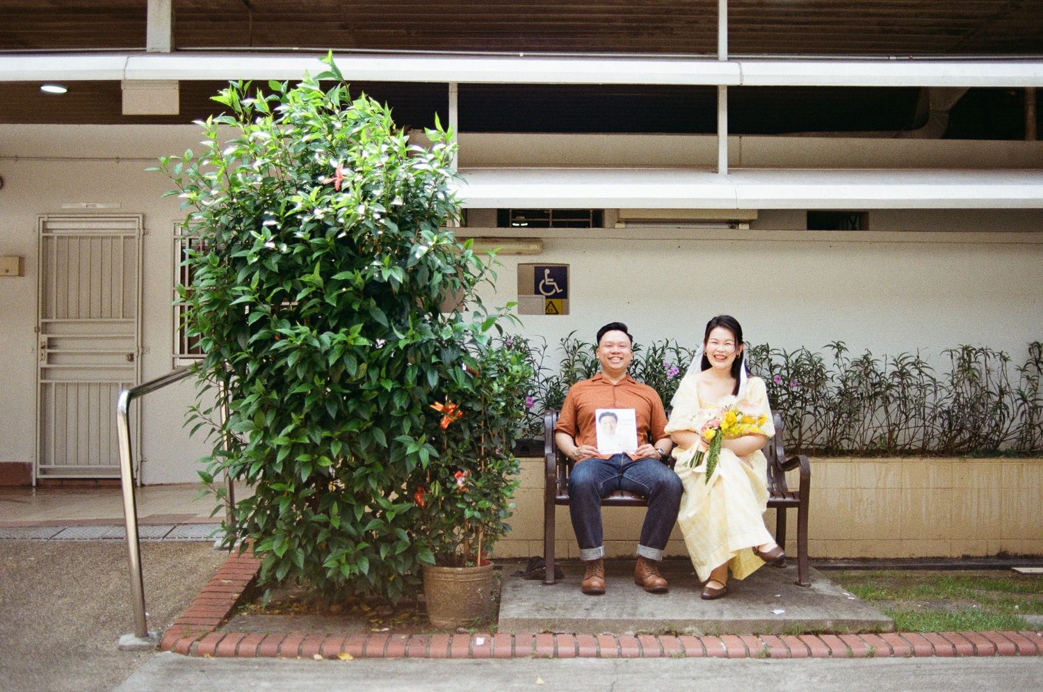 A couple relaxes on a bench beside lush green plants and brick pavers in a courtyard setting.