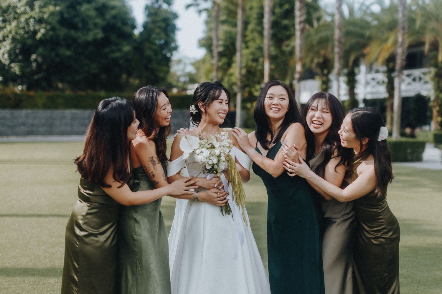 Wedding party in olive green bridesmaid dresses and white bridal gown share joyful moment outdoors on sunny day.
