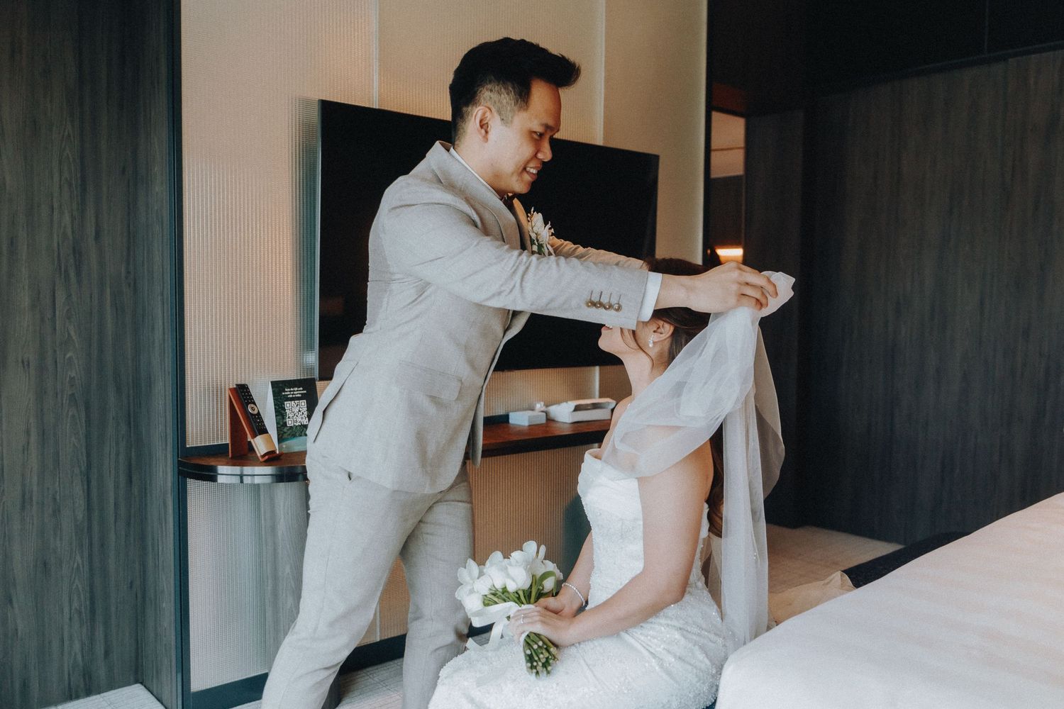 A tender moment between newlyweds in a hotel room with the groom in beige suit helping the bride.