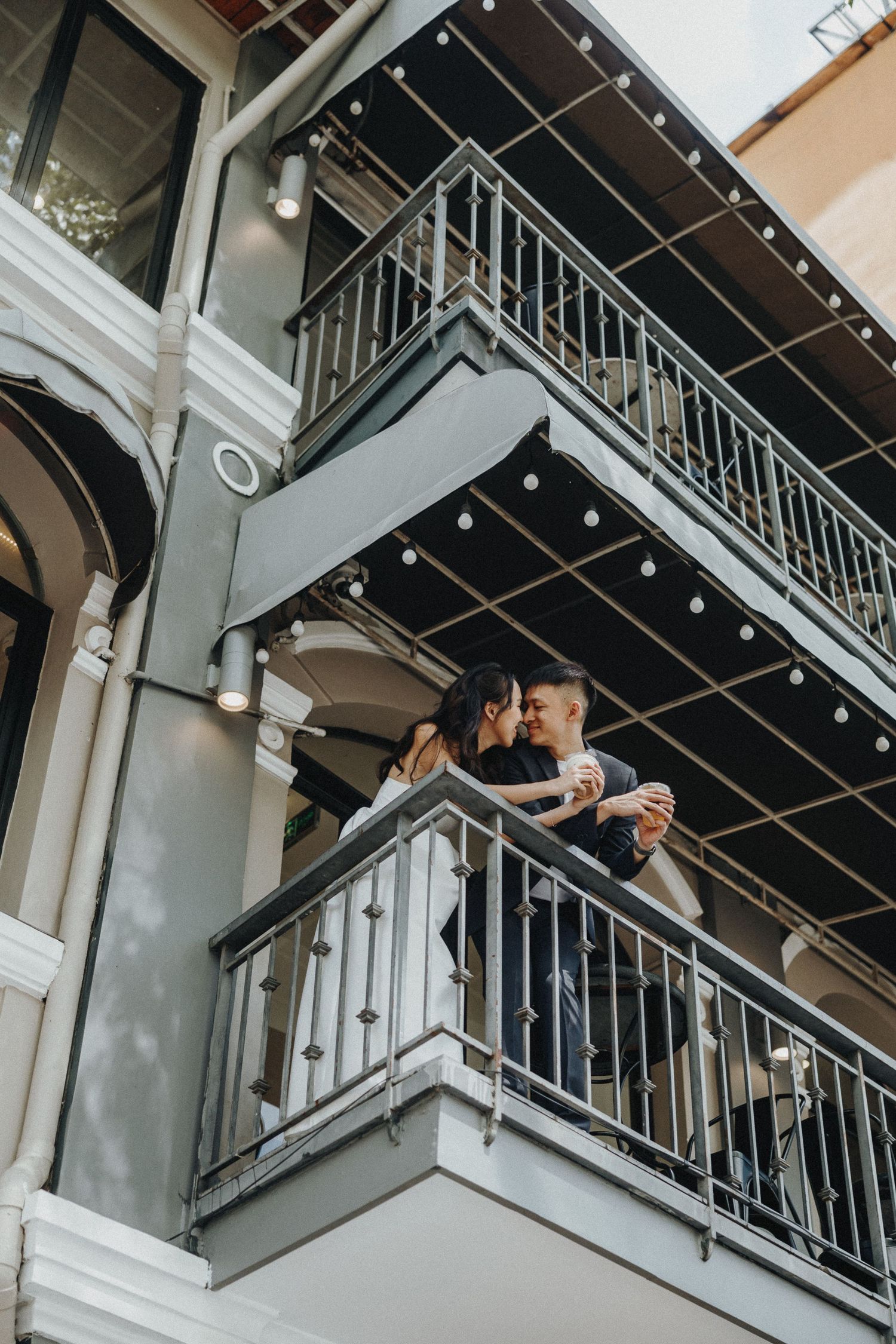 A couple holds champagne glasses and toasts on a modern urban balcony with metal railings.