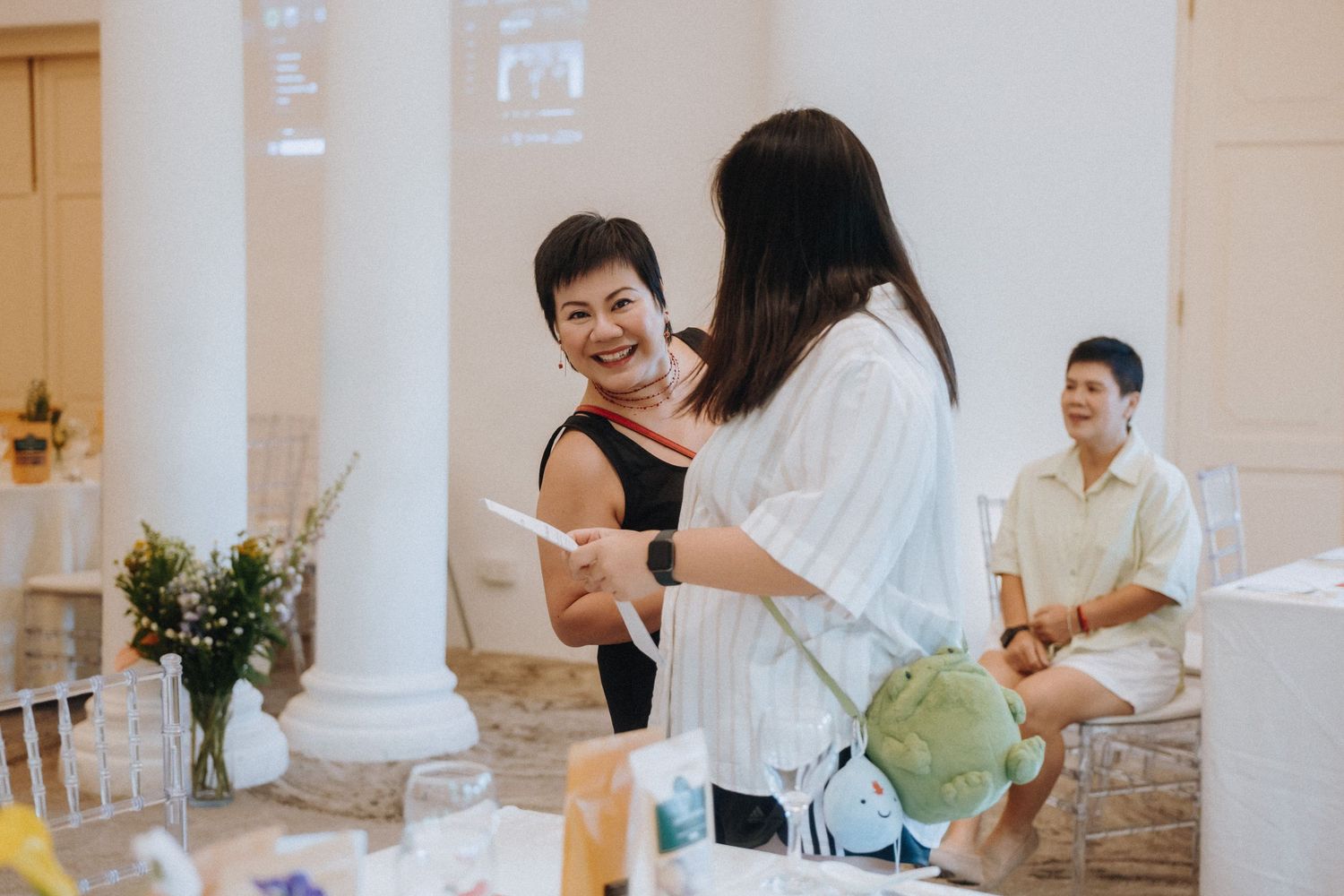 Two women smile and chat at an elegant wedding reception venue with white columns and floral arrangements.