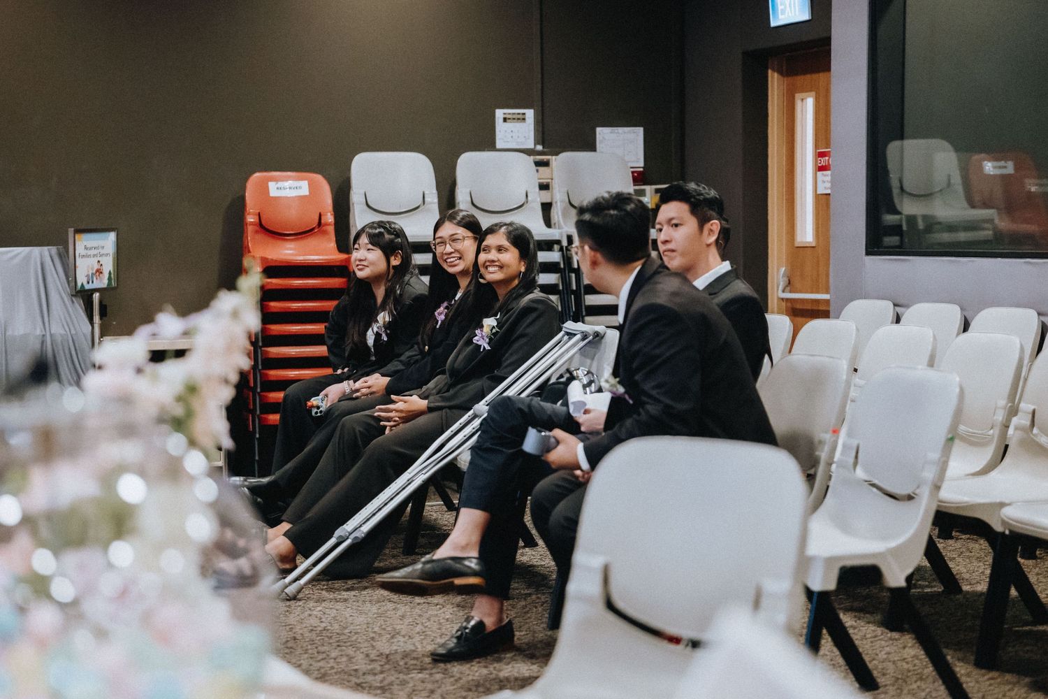 Wedding guests seated in white chairs during a ceremony with orange stacked chairs visible in background.