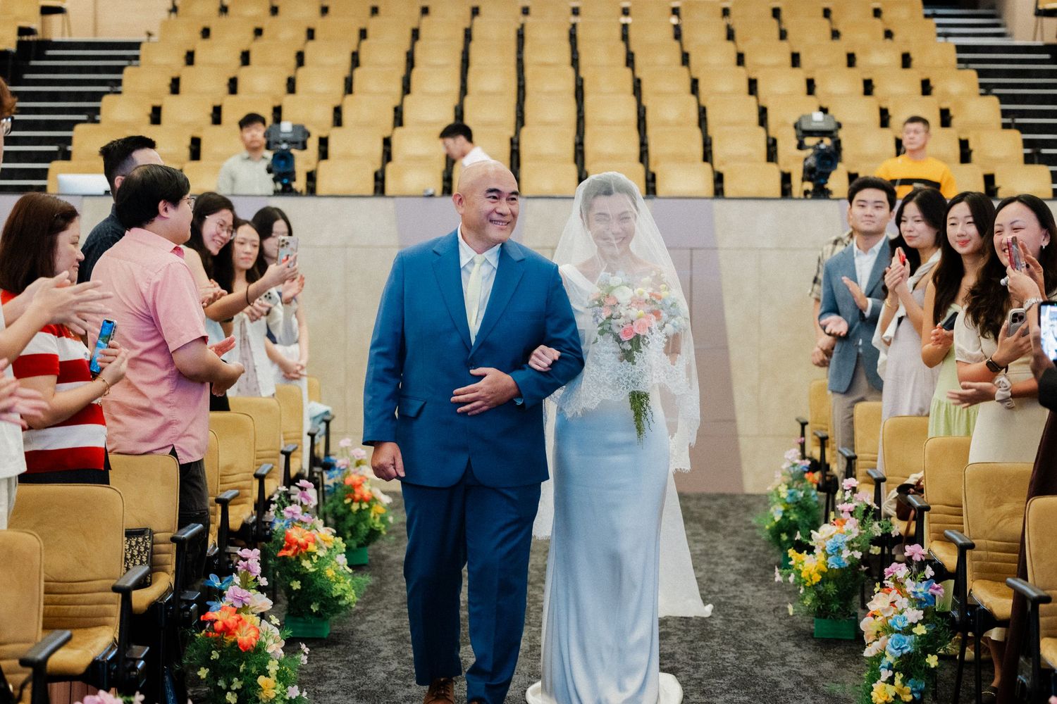 Bride in blue gown walks down aisle with older man in blue suit at wedding ceremony.