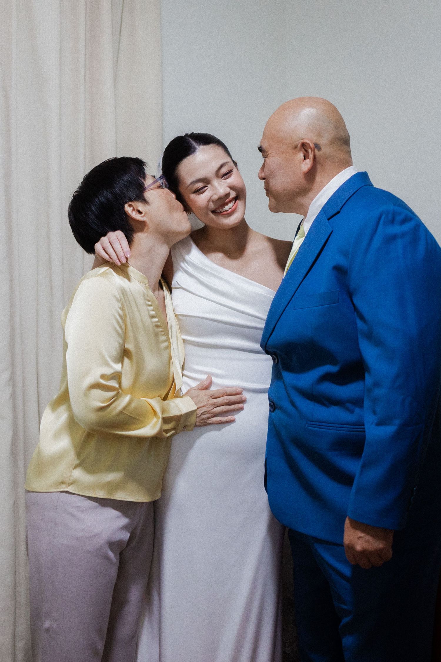 Bride smiling joyfully as her parents kiss her cheeks in a tender wedding day moment.