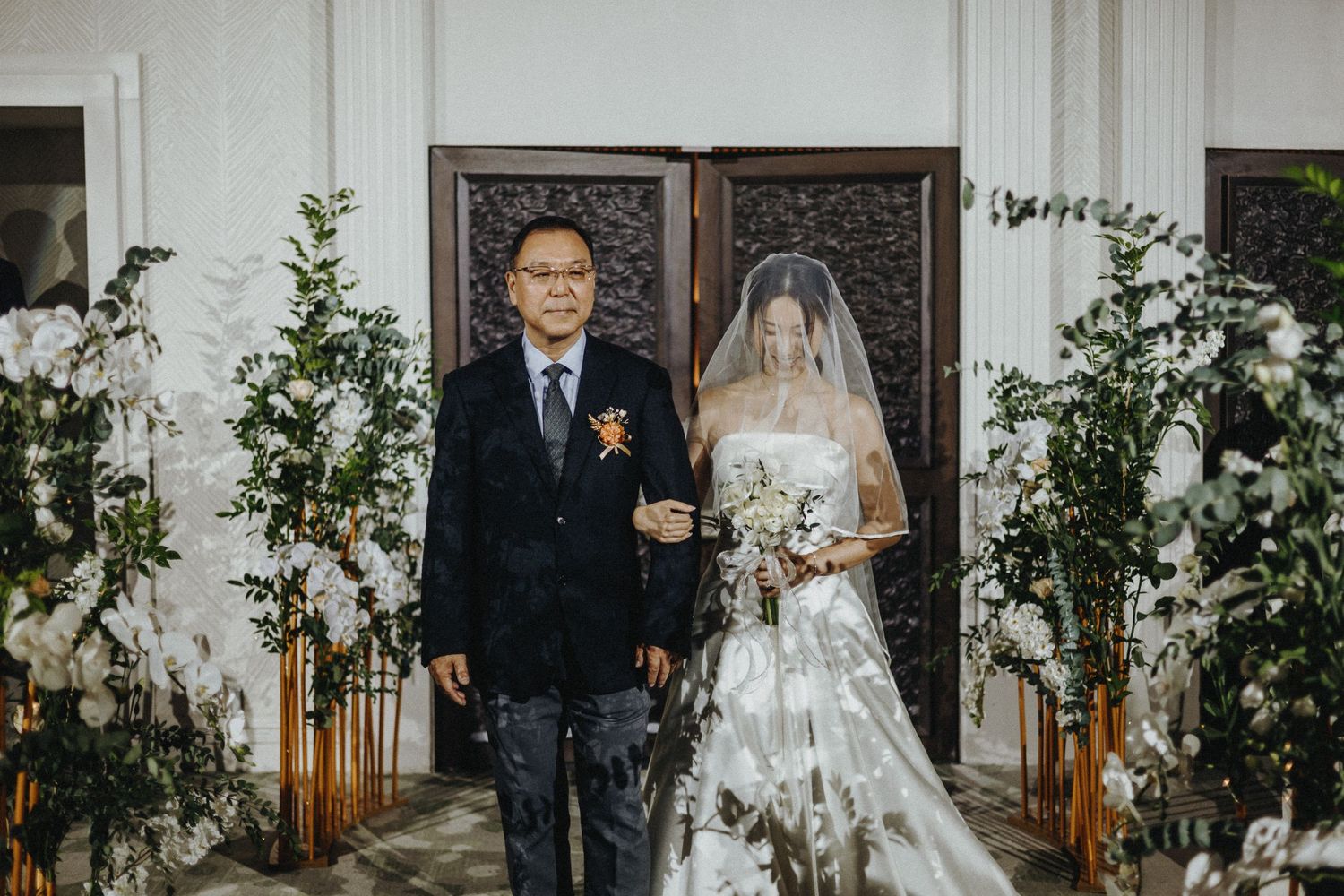 A formal wedding ceremony takes place in an elegant venue decorated with greenery and wooden screens.
