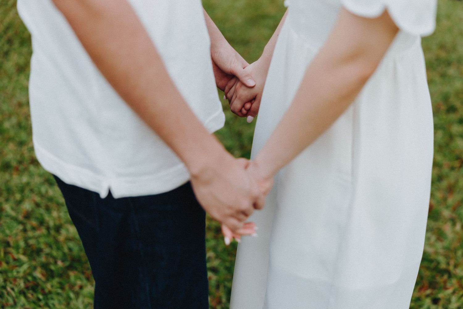 A couple in white and black formal attire holds hands tenderly in a lush garden setting.