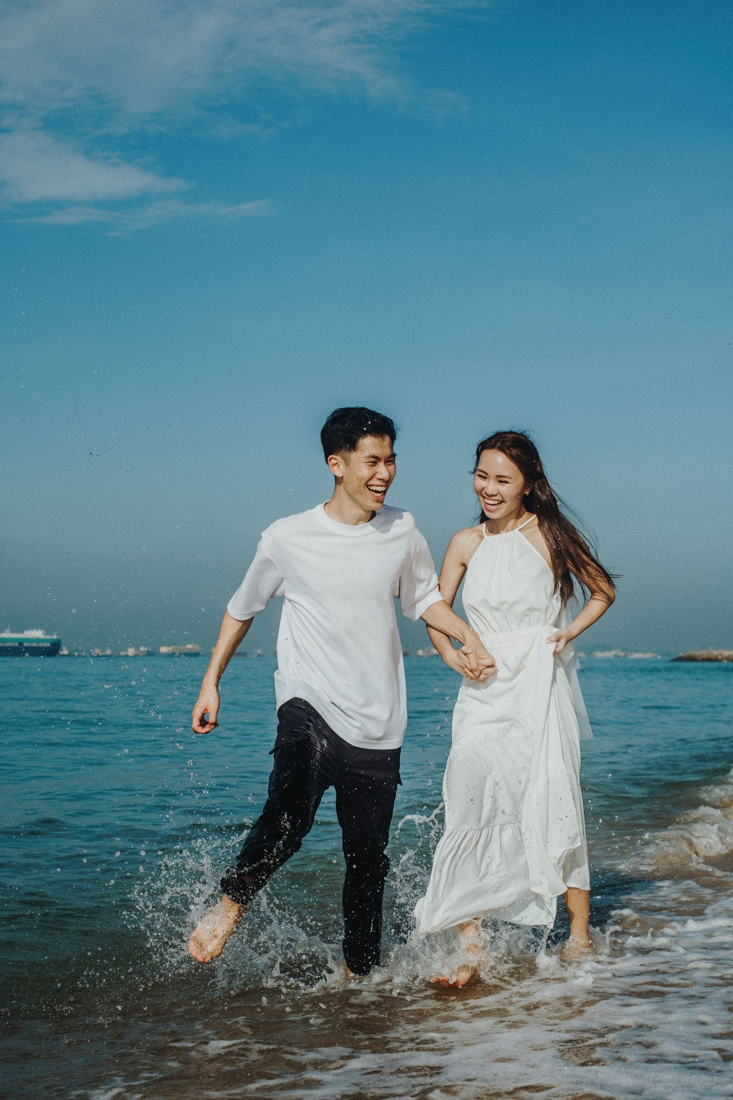 A playful couple runs through ocean waves wearing white outfits on a sunny beach day.