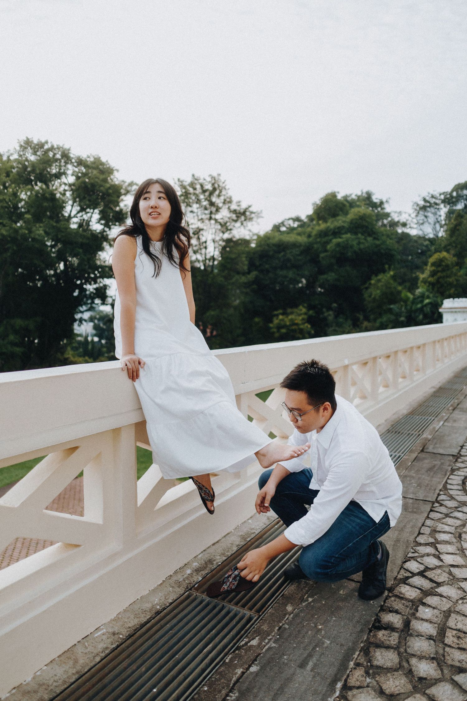 A couple creates a playful moment on a stone bridge with trees in the background on a cloudy day.