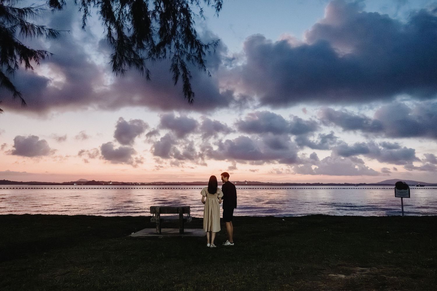 Silhouetted couple stands by a bench watching dramatic sunset over ocean with cloudy sky and palm trees.