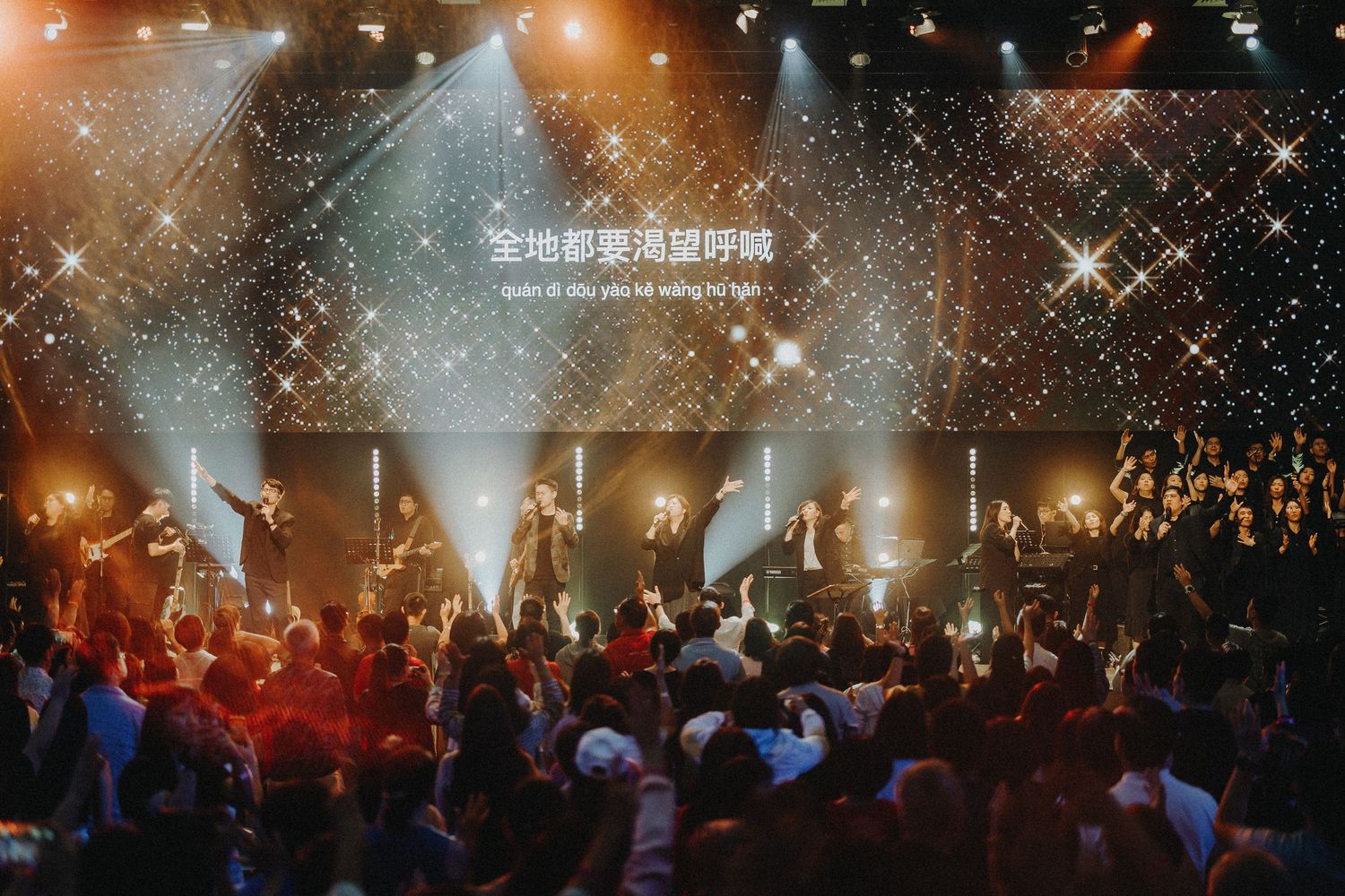 A concert venue filled with warm orange lighting and silhouetted crowd under sparkling light effects and fog.