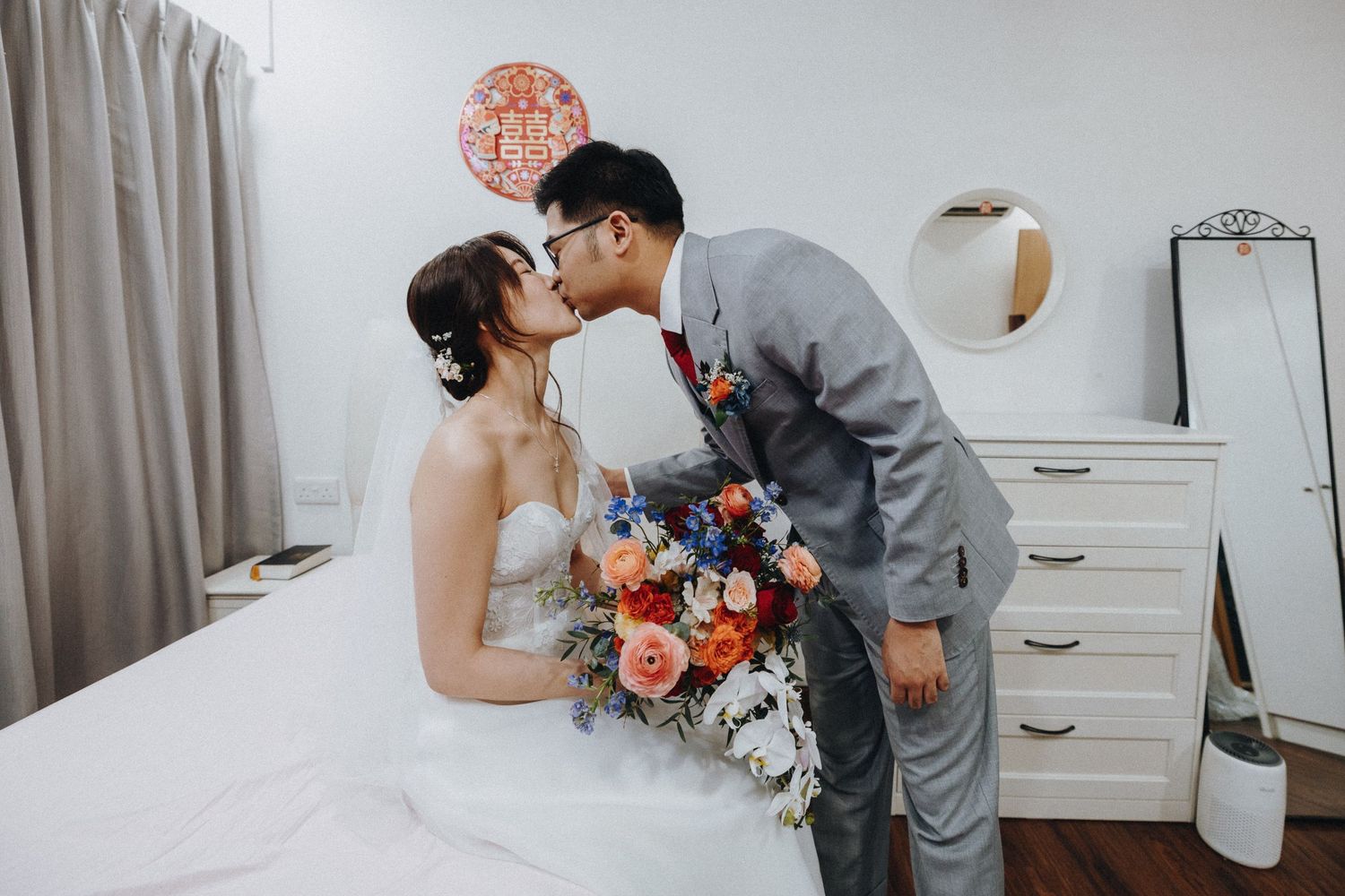 A romantic wedding couple sharing intimate moments while holding coral and peach floral bouquets in a light-filled room.