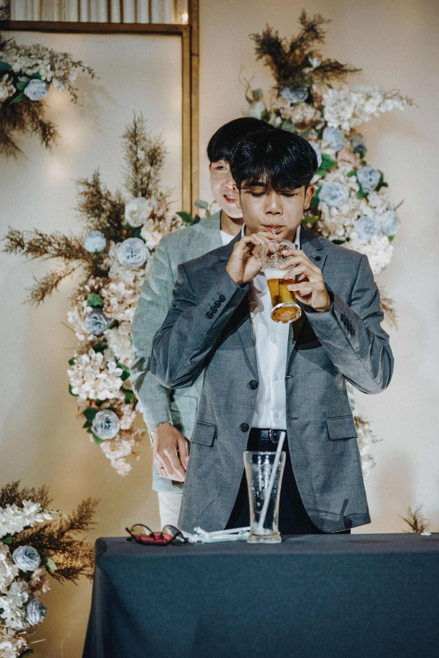 Wedding guest in grey suit enjoying a drink at decorated reception table with blue floral arrangements.