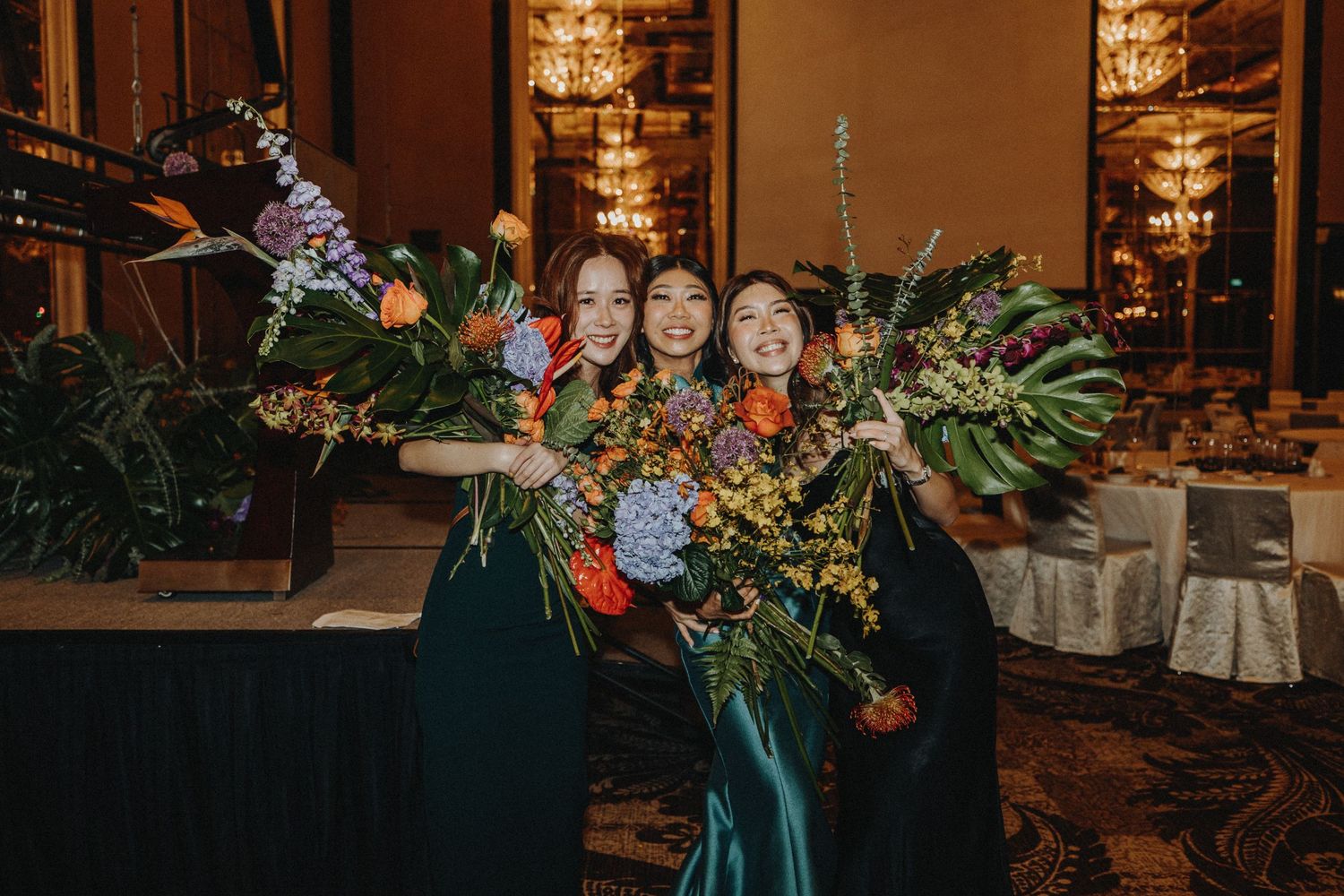 Bridesmaids in dark gowns posing with tropical flower arrangements at wedding reception.