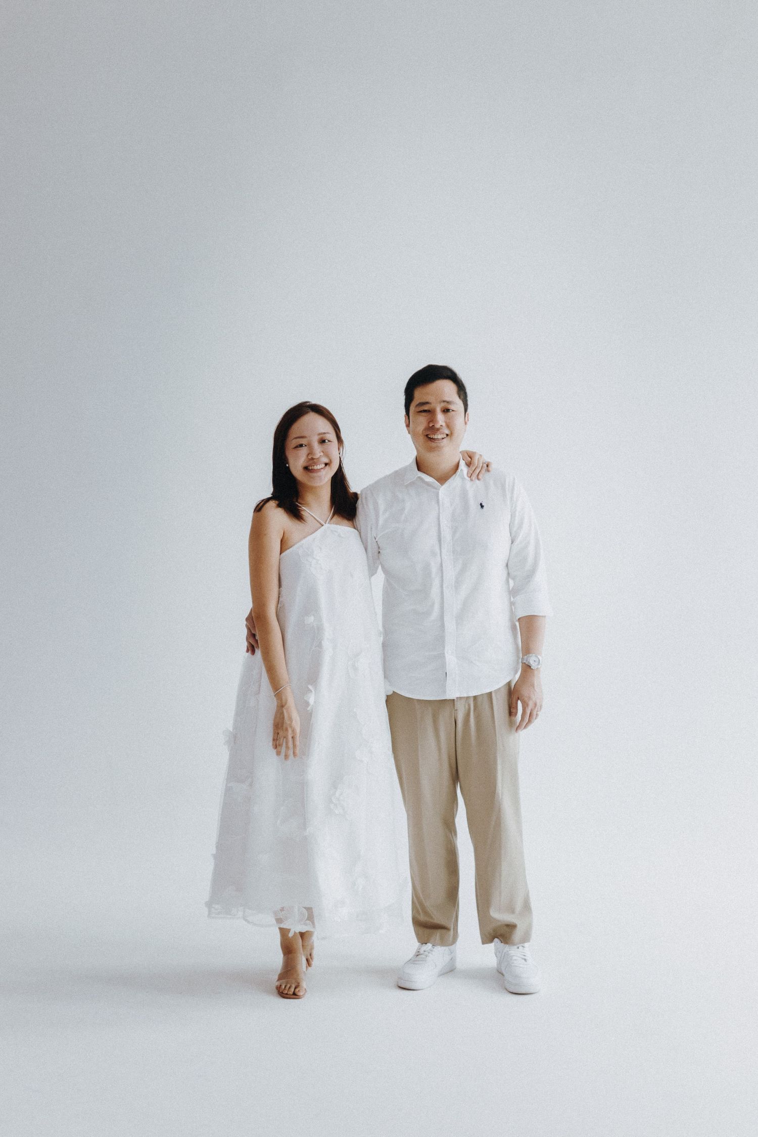 A couple in white outfits poses against a minimalist grey backdrop for an elegant portrait session.